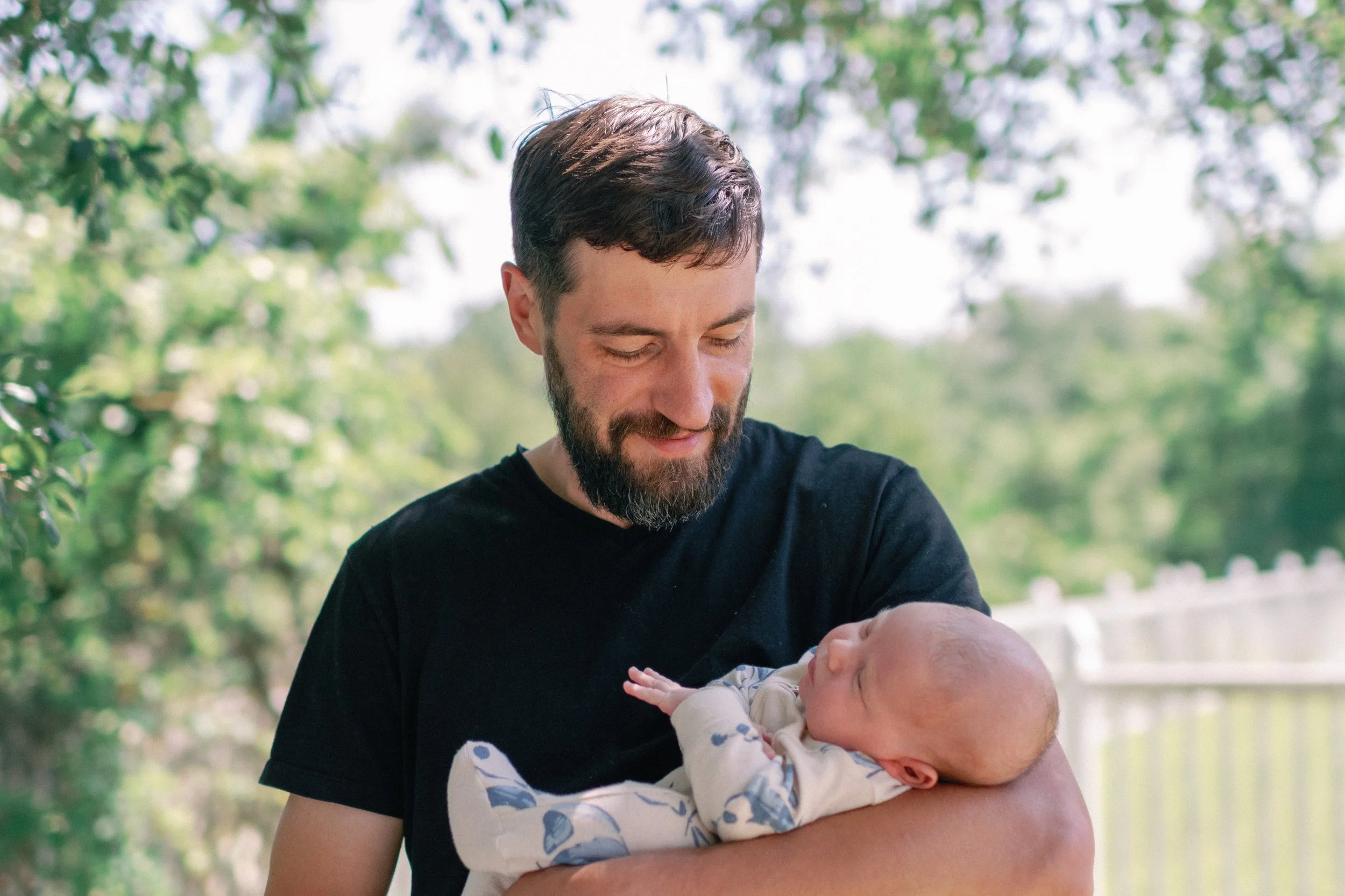 A man with a beard and short hair holding a newborn baby outdoors with green trees and a white fence in the background.