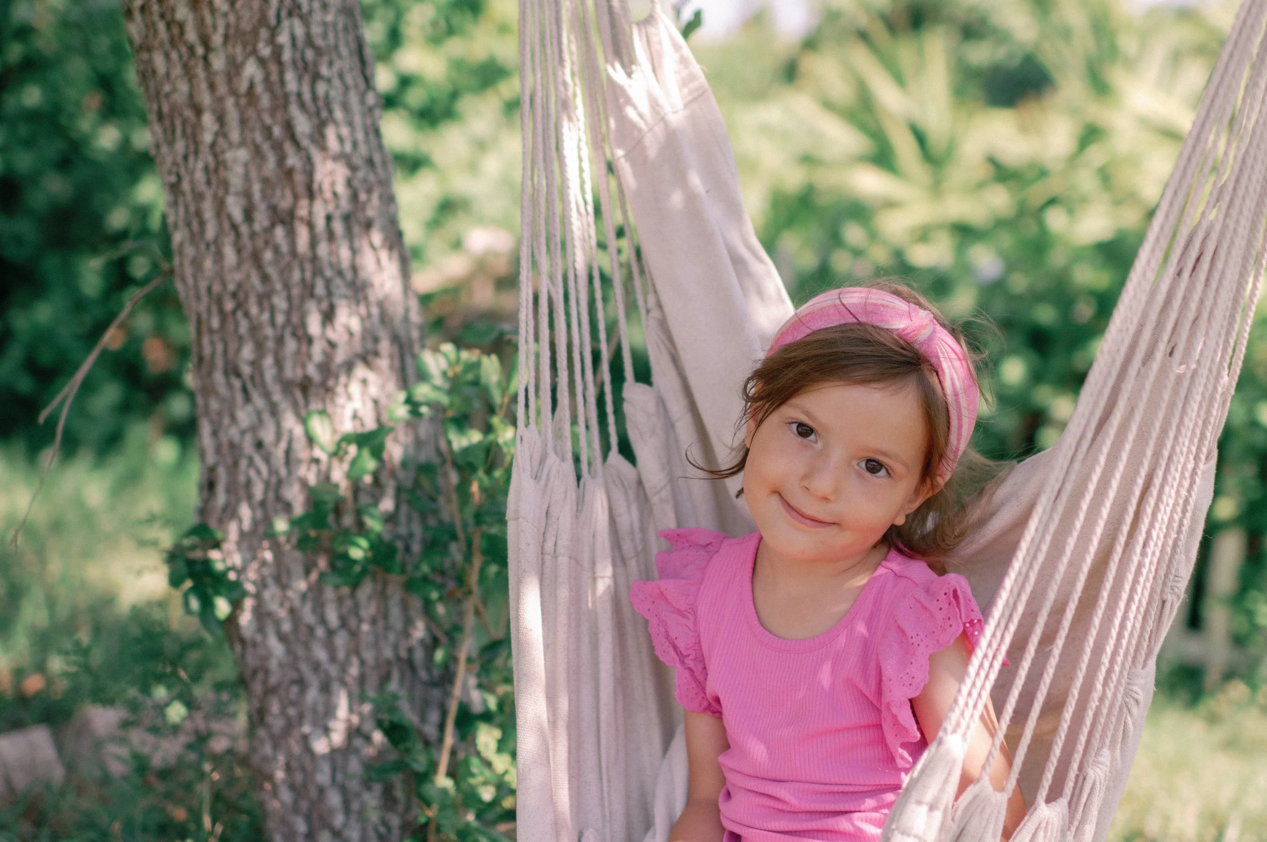 A young girl with brown hair wearing a pink headband and pink dress, sitting on a hammock outdoors in a green garden.