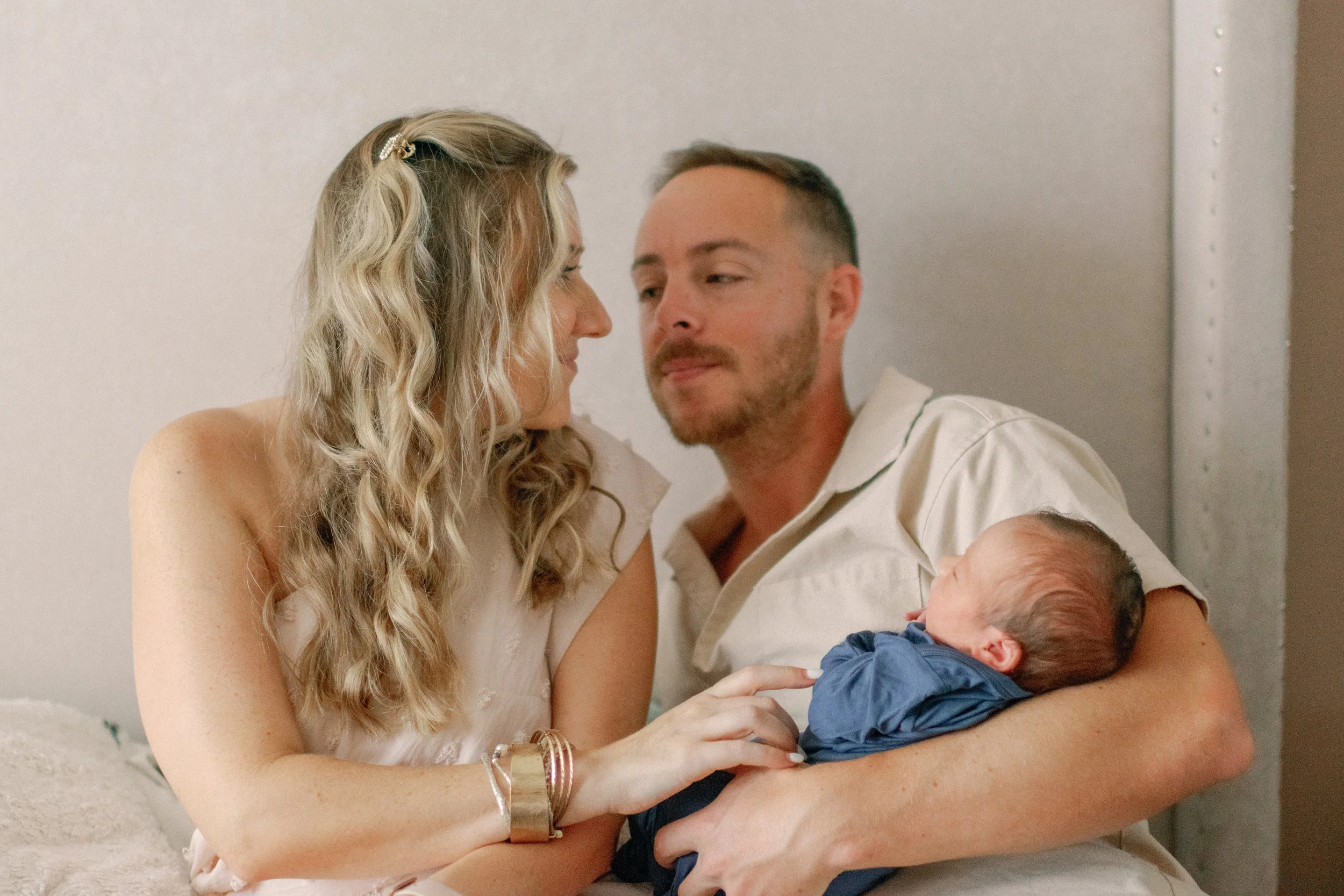A couple with a newborn baby in a cozy indoor setting. The woman with blonde, wavy hair and jewelry is sitting close to the man with short hair and a beard, who is holding the baby in his arms. The baby is dressed in blue and appears to be asleep.
