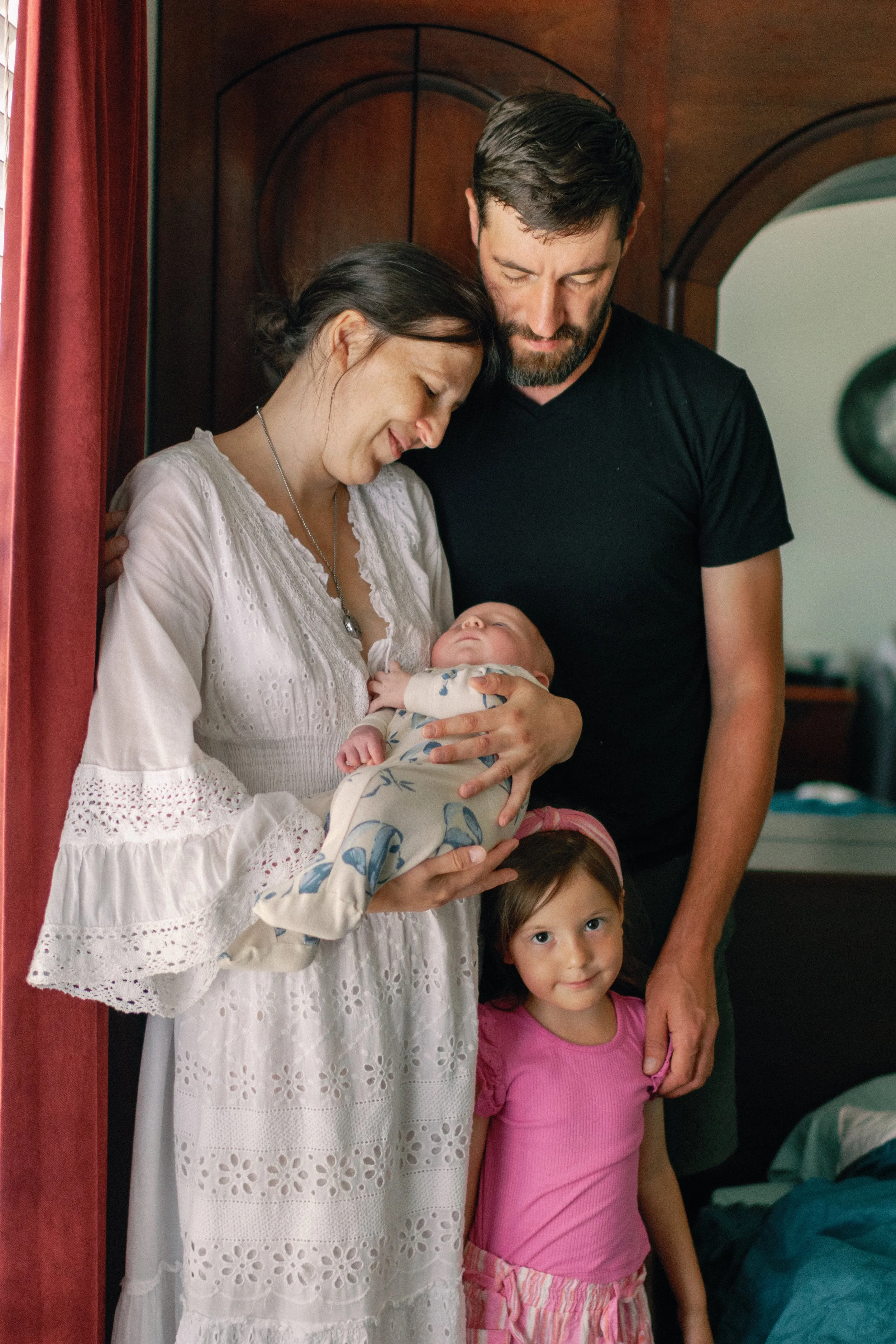 Family of four, including a woman holding a baby, a man, and a young girl, inside a home, smiling and looking at the baby.