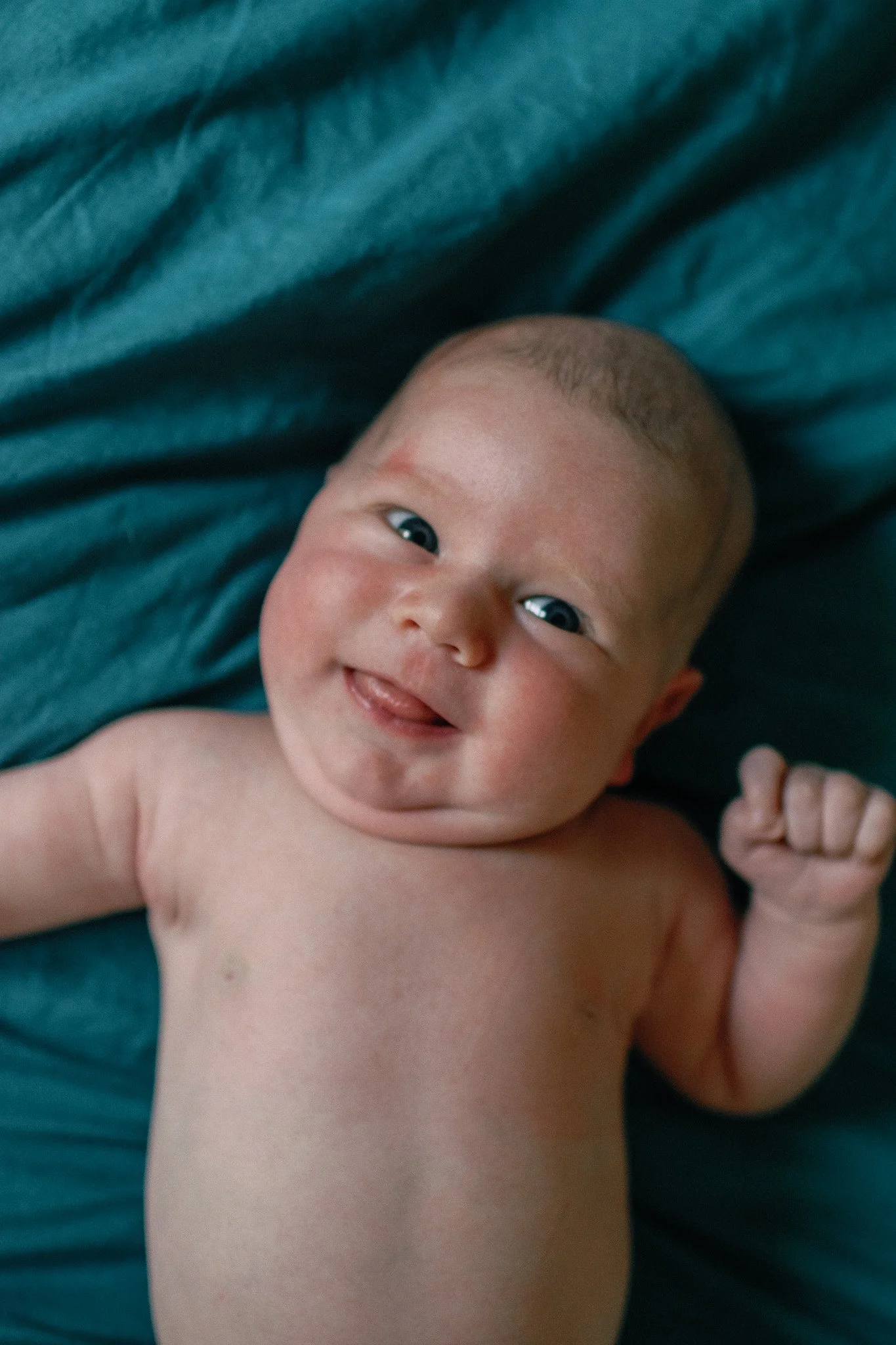 Smiling baby lying on a teal blanket, showing a happy expression and a small fist raised.