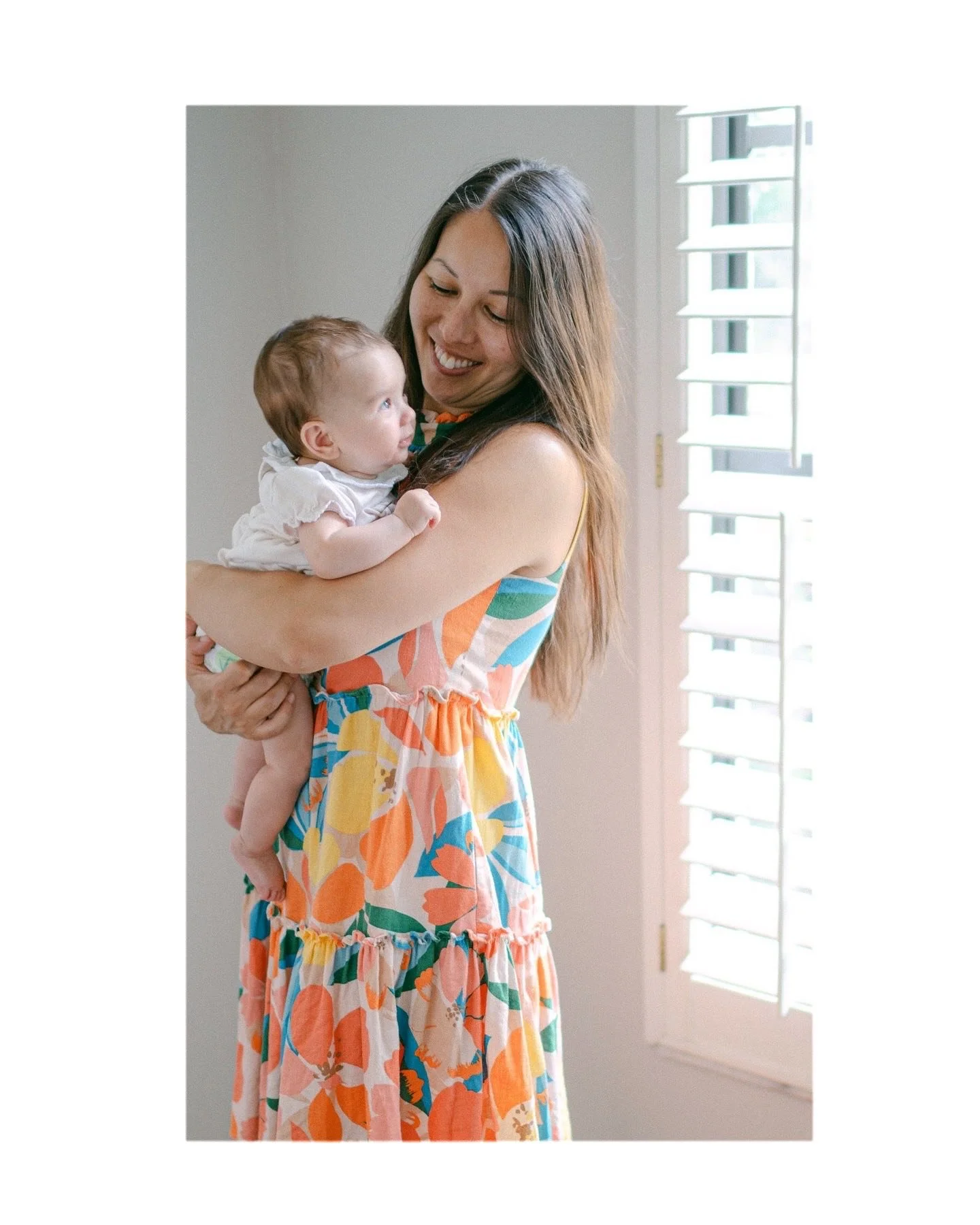 A woman holding a baby indoors next to a window with white shutters, both looking at each other.