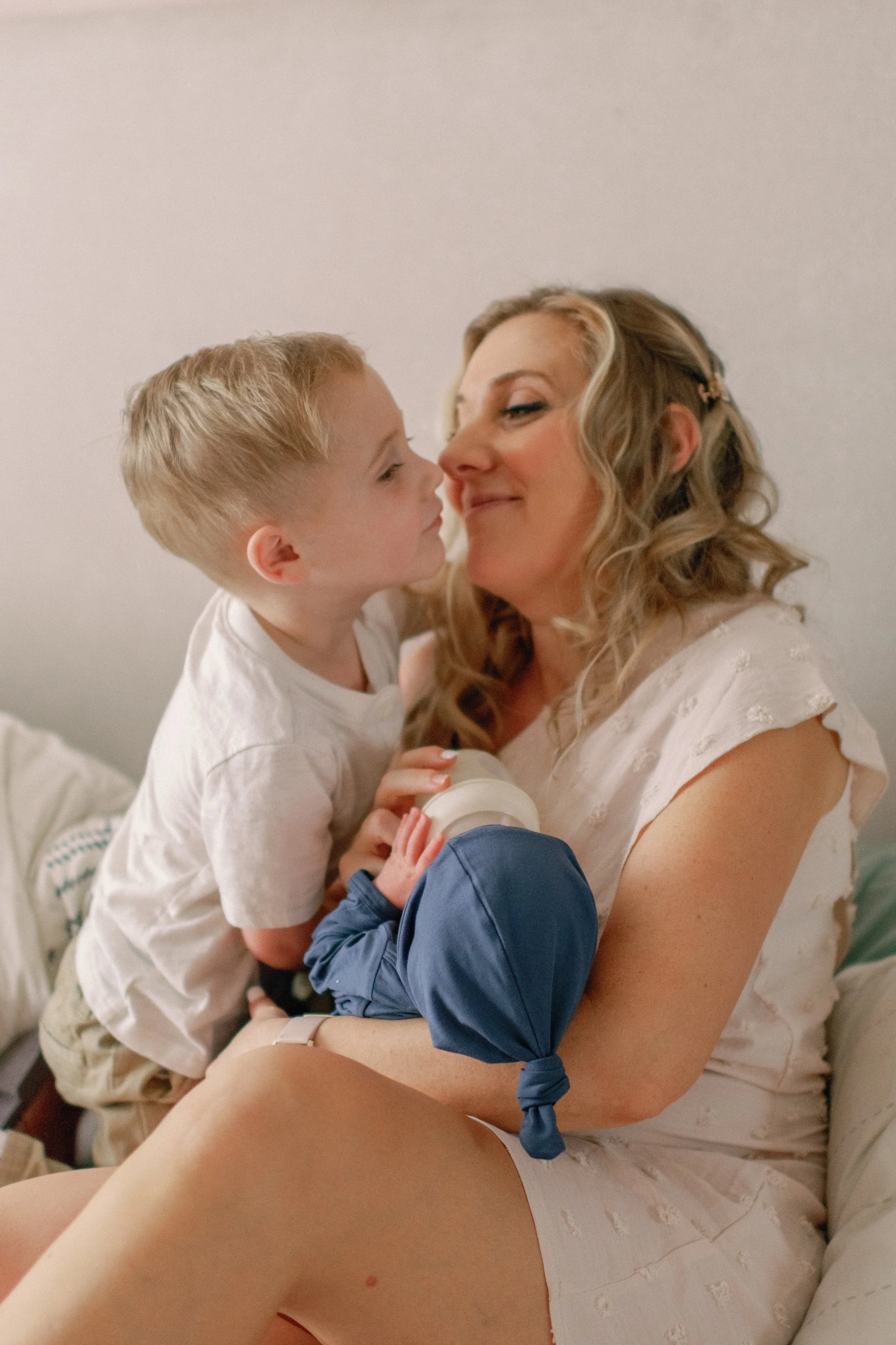A woman with makeup and curly blonde hair interacts lovingly with a young boy, holding a baby bottle. They are seated on a bed with neutral-colored bedding.