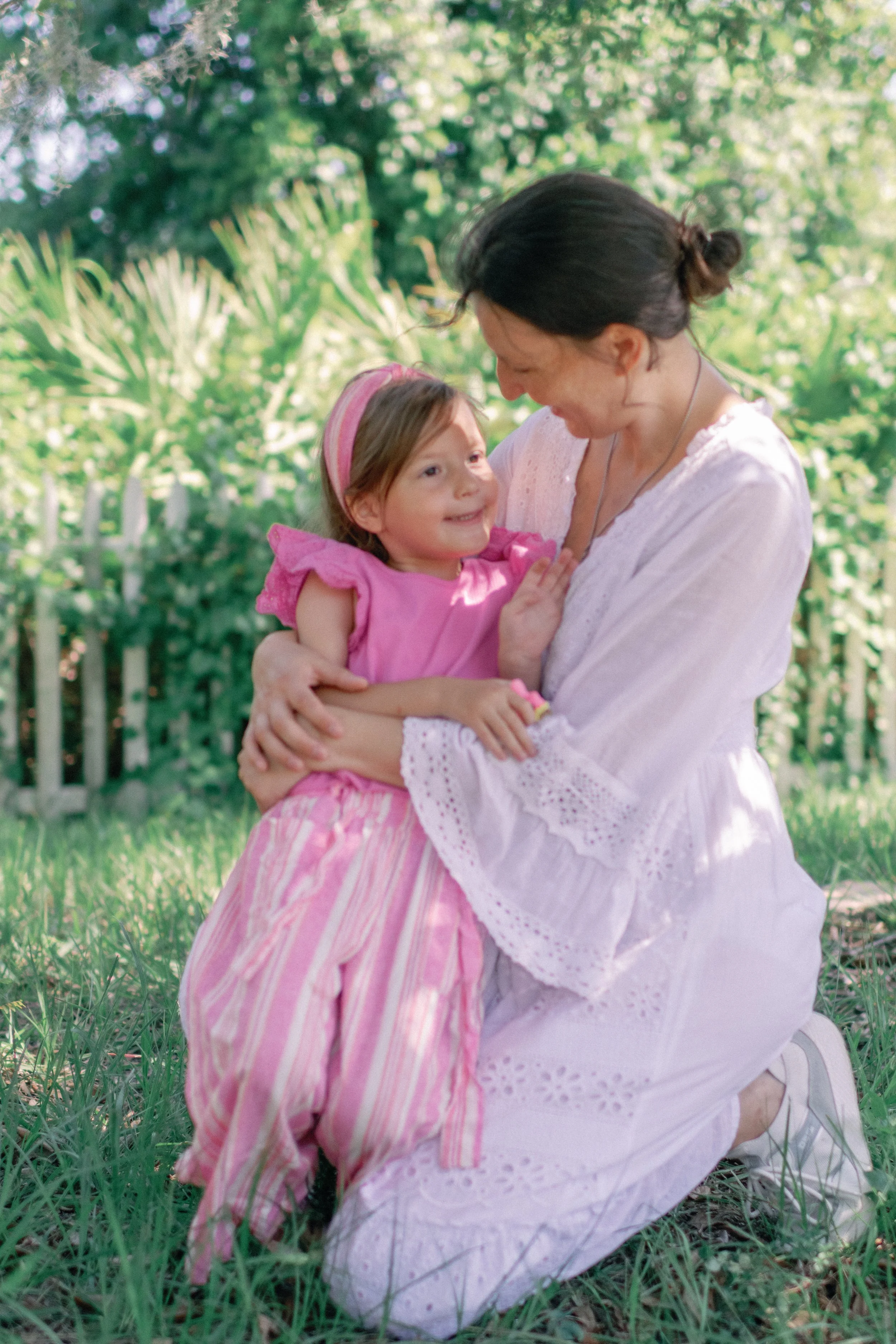 A woman kneeling on the grass, hugging a young girl in a garden with trees and a white fence in the background.
