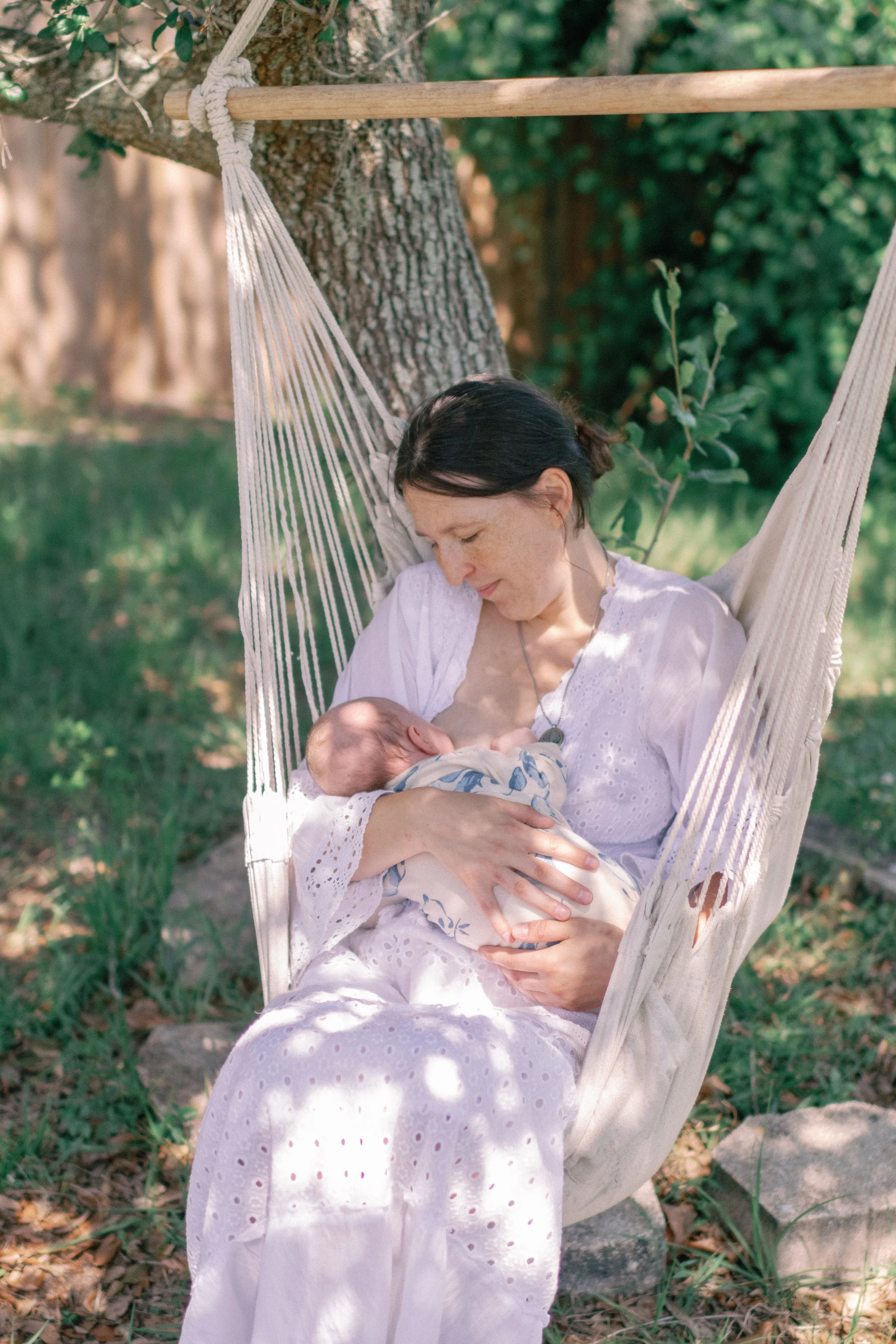 Woman sitting in a hammock under a tree, breastfeeding a baby outdoors in a garden.