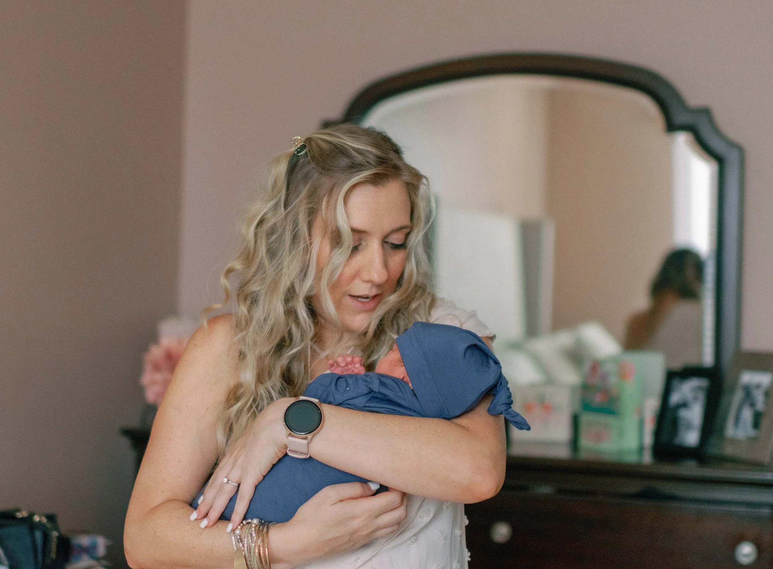 A woman holding a newborn baby in a room with a mirror and framed pictures on a dresser.