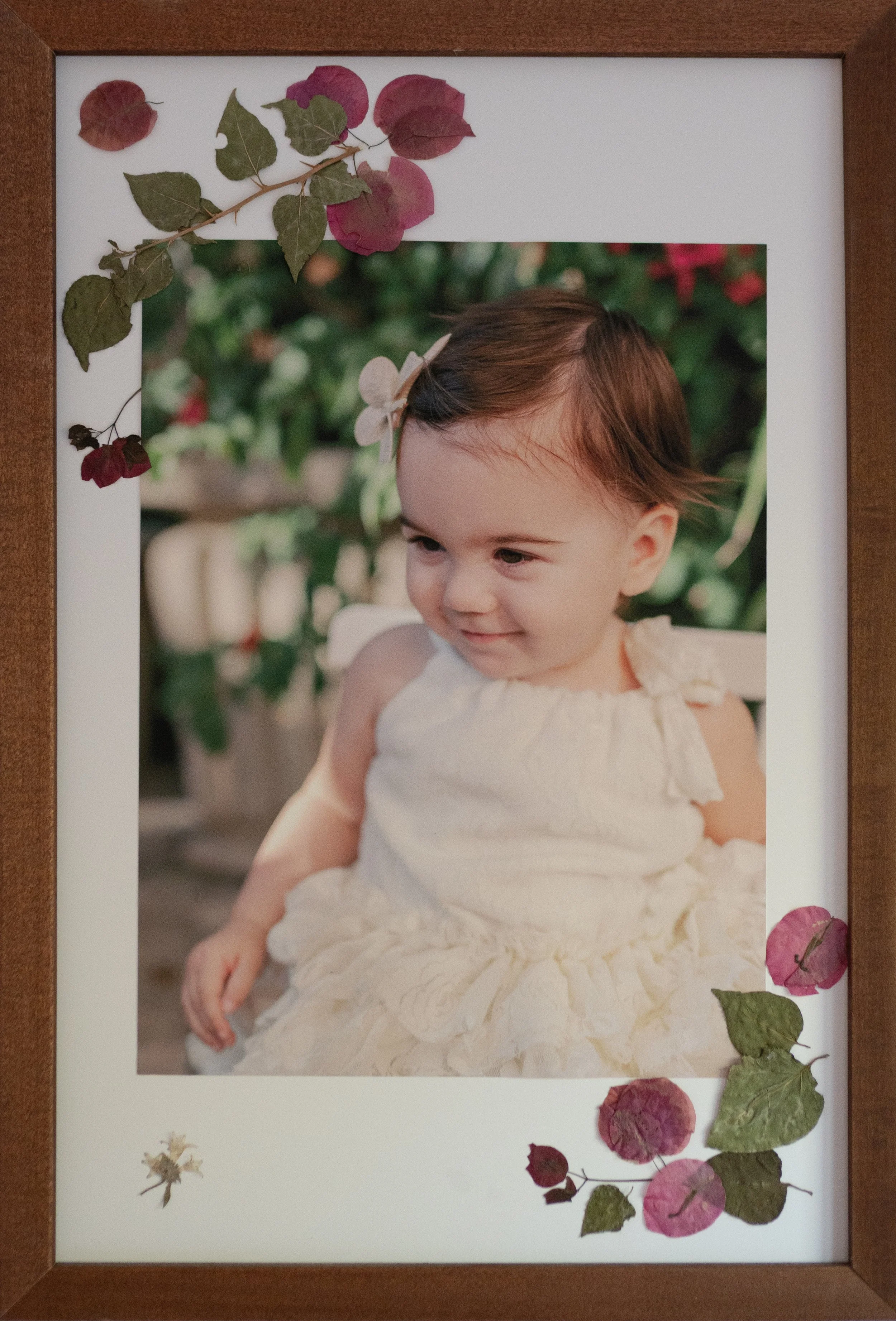 A framed photograph of a young girl with short brown hair, wearing a cream-colored dress with ruffled sleeves, sitting outdoors with greenery and pink flowers in the background. The frame is decorated with dried leaves and pink flowers around the edges.