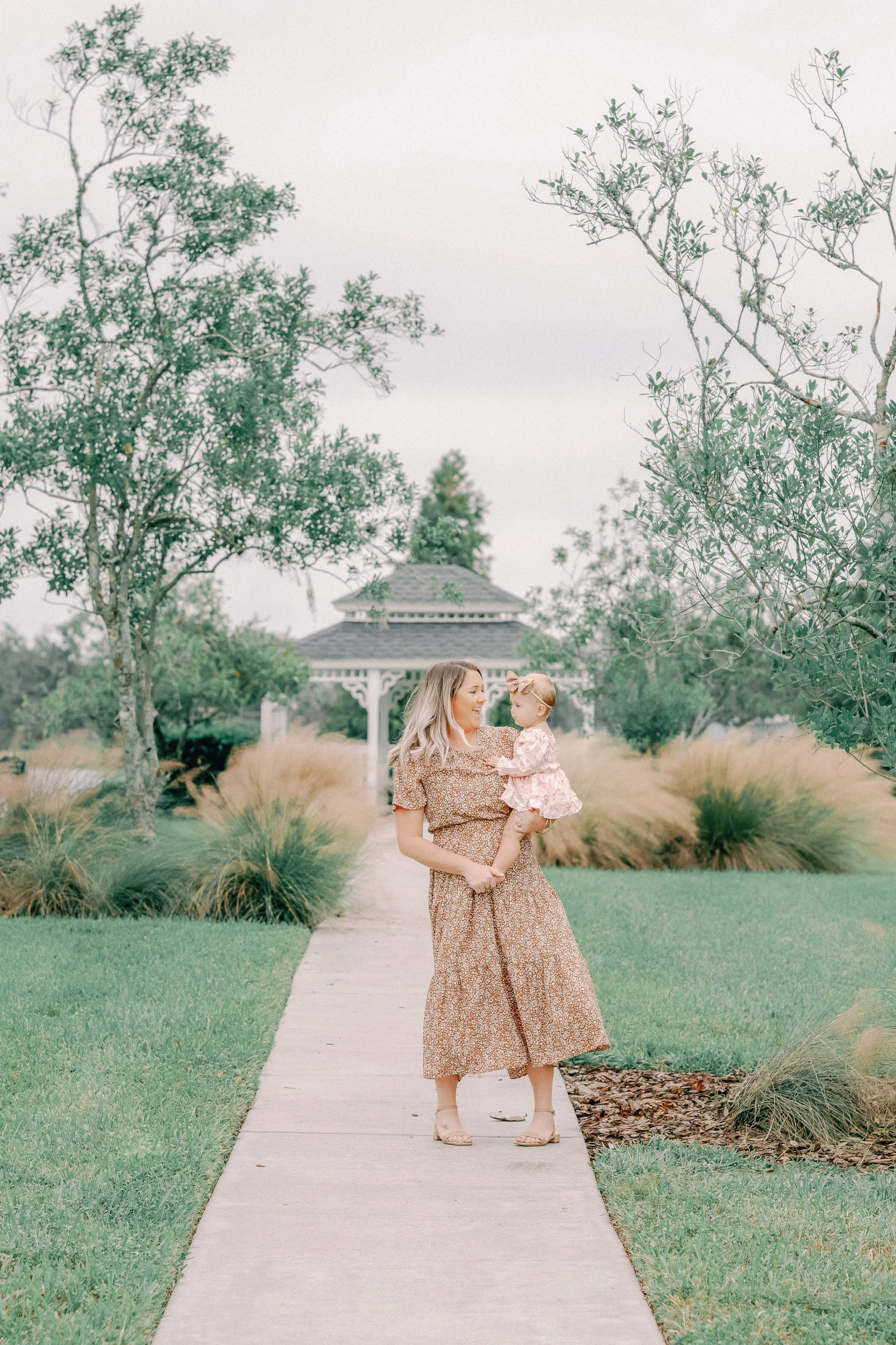 A woman holding a young girl on a sidewalk in a park with grass and trees, a gazebo in the background, and cloudy sky.