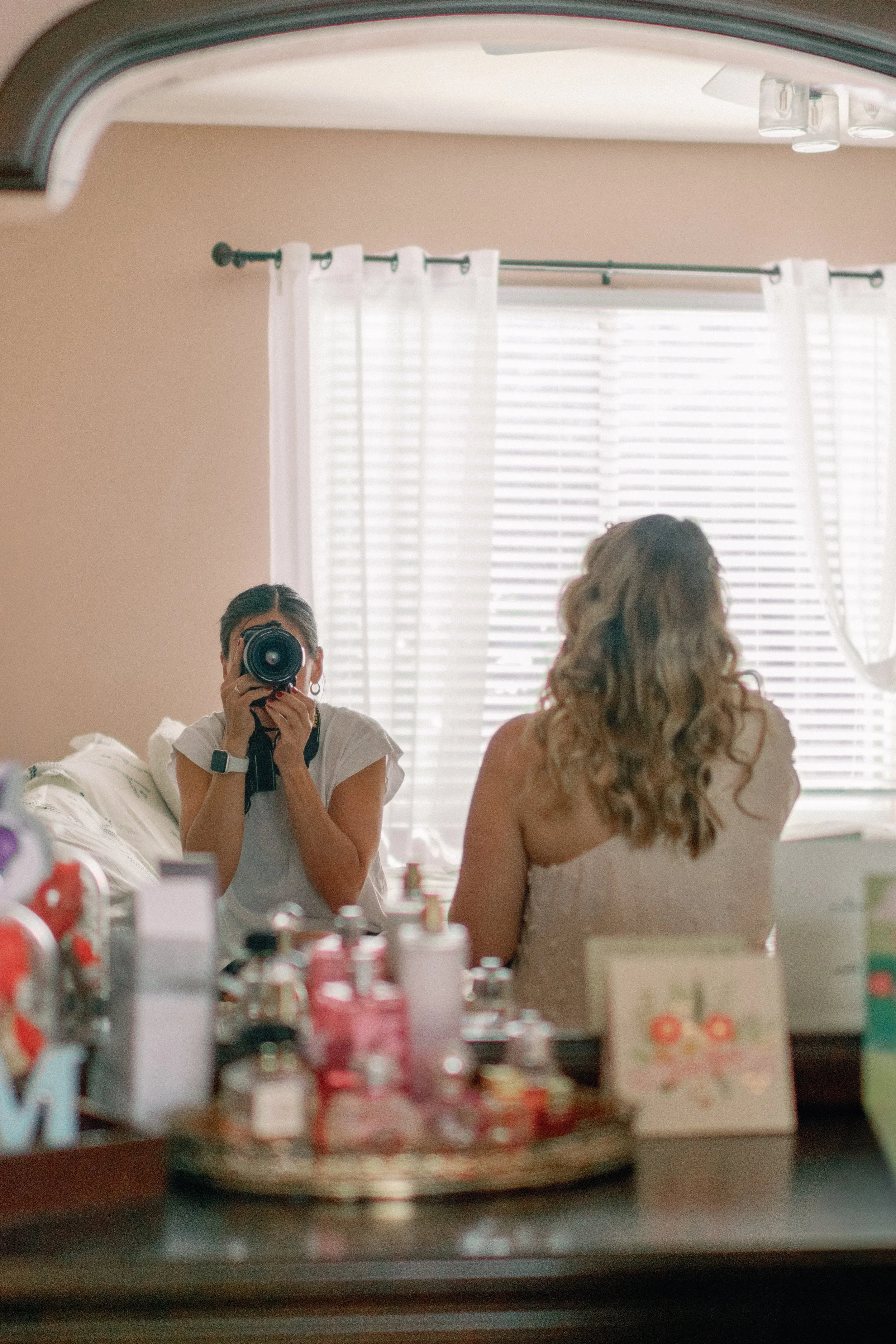 A woman with curly hair sitting in front of a mirror with her back to the camera, facing a window with blinds and white curtains. Another woman is taking her photo with a camera, reflected in the mirror. The surface below the mirror has various bottles and a card with a floral design.