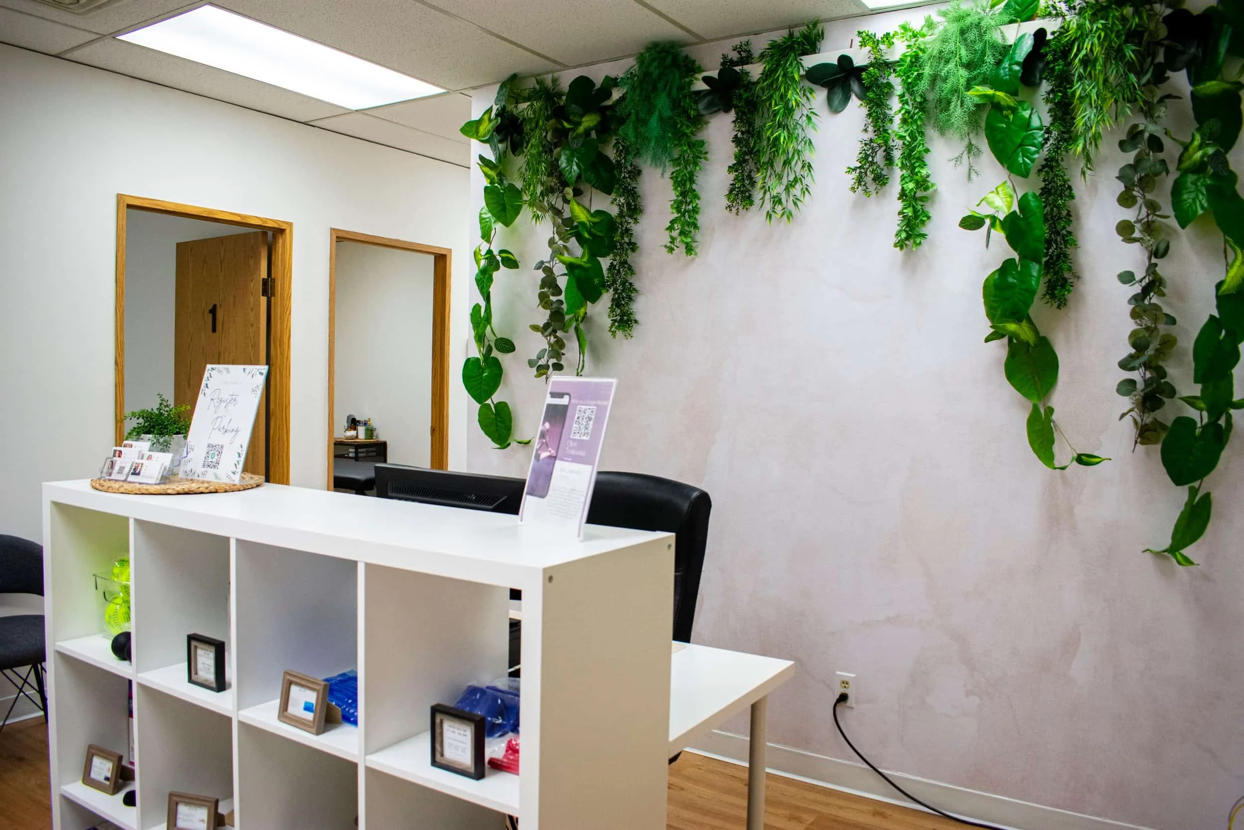 Reception area with a white desk, shelving, green plants, and touches of ce sport therapy; wooden doors and chairs in the background.