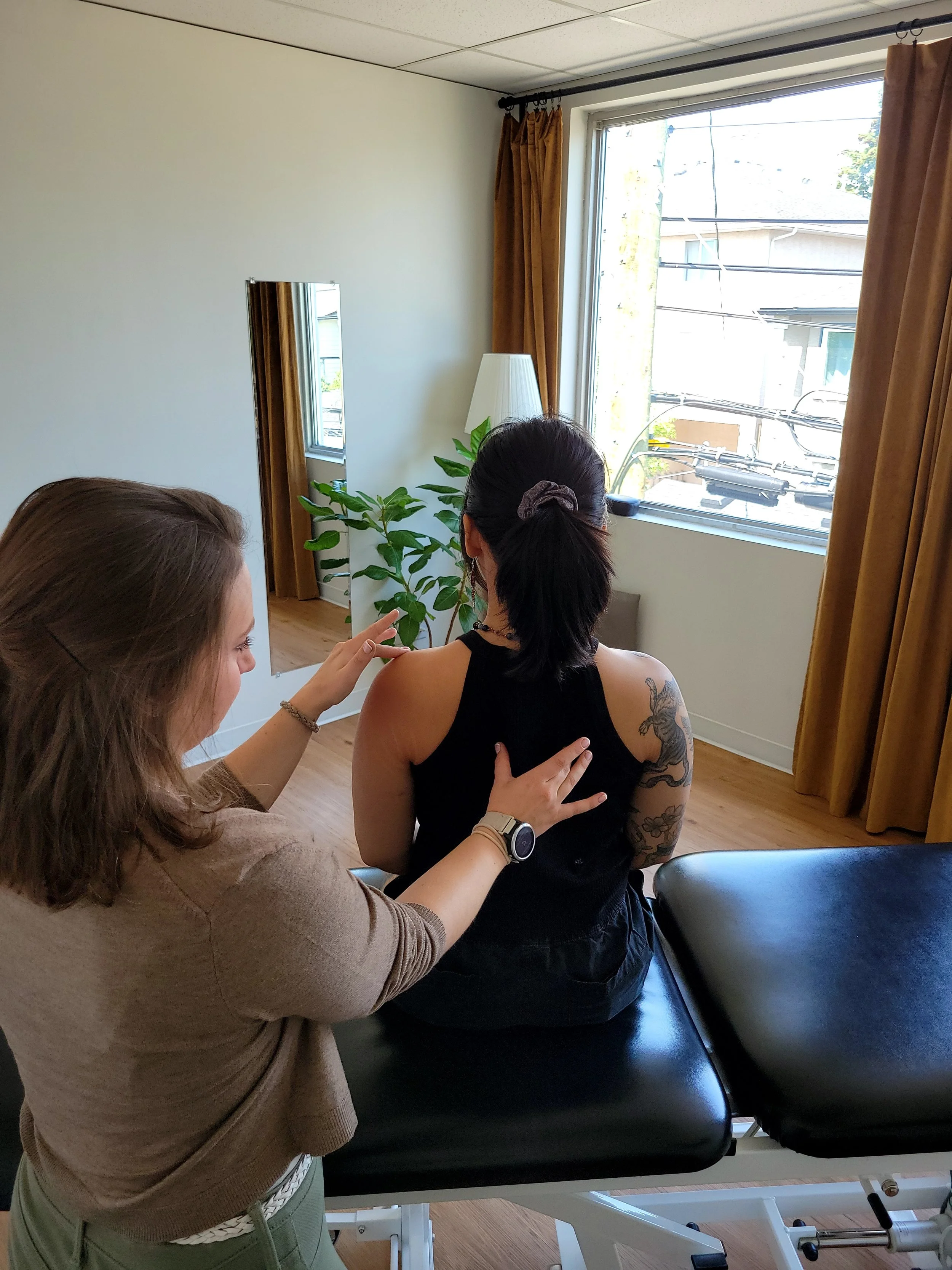 A woman with dark hair in a ponytail and tattoos on her right arm is sitting on a medical examination table wearing a black sleeveless top. Another woman with shoulder-length brown hair, wearing a beige sweater, is standing beside her, touching her s