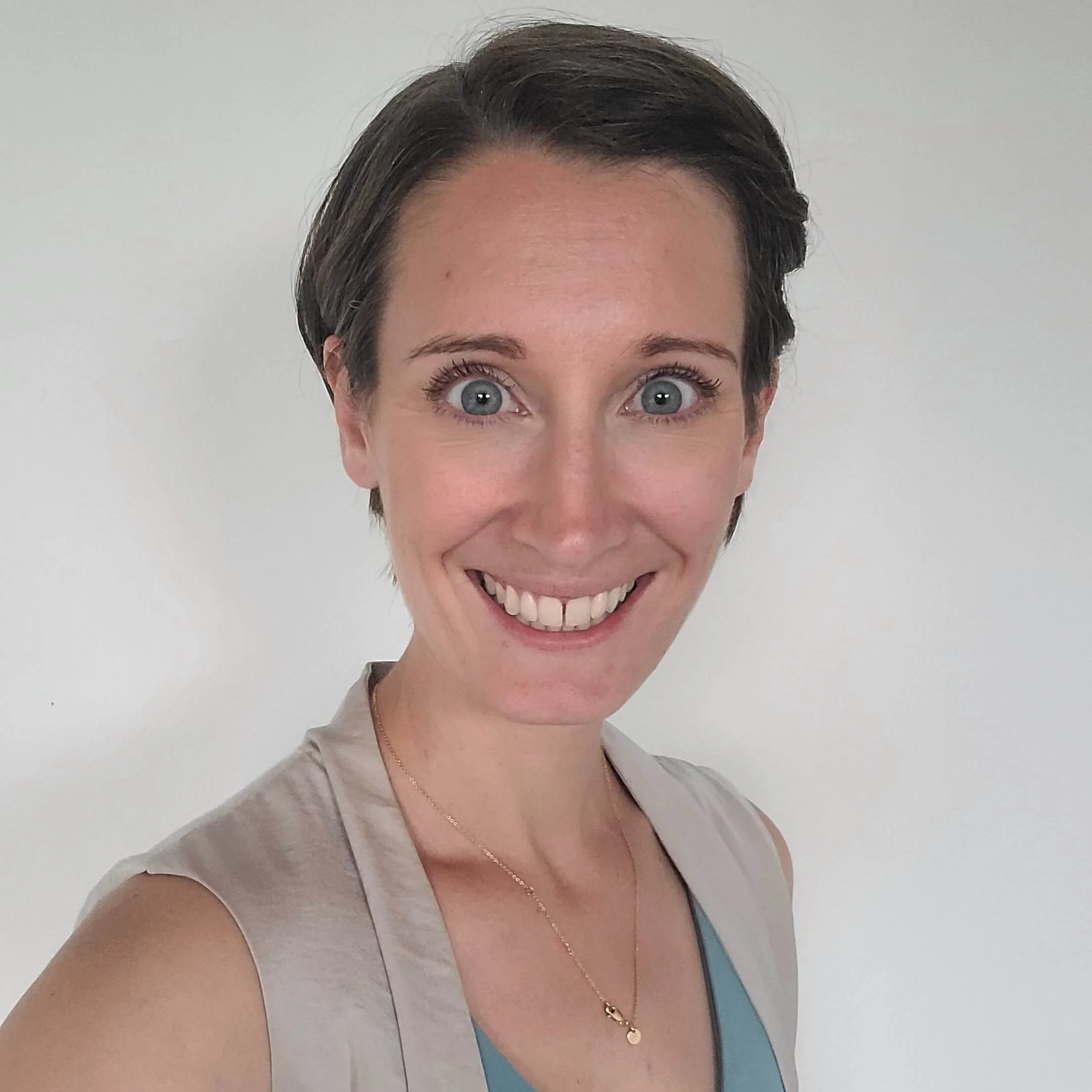 Woman with short brown hair and blue eyes smiles, looking relieved from back pain, in a light vest and blue top against a white background.