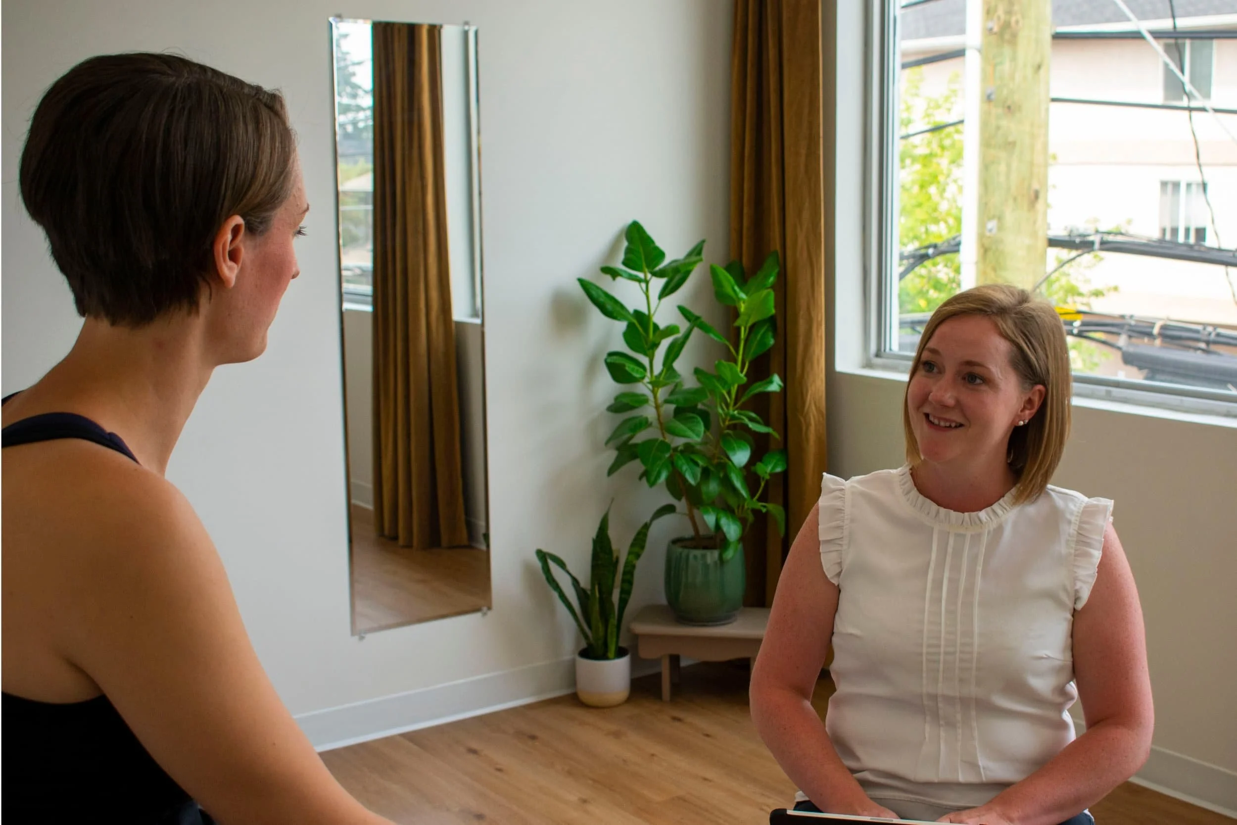 Two women discuss athletic therapy in a bright room with wooden floors, large windows, mirrors, and plants—focusing on rotator cuff pain.