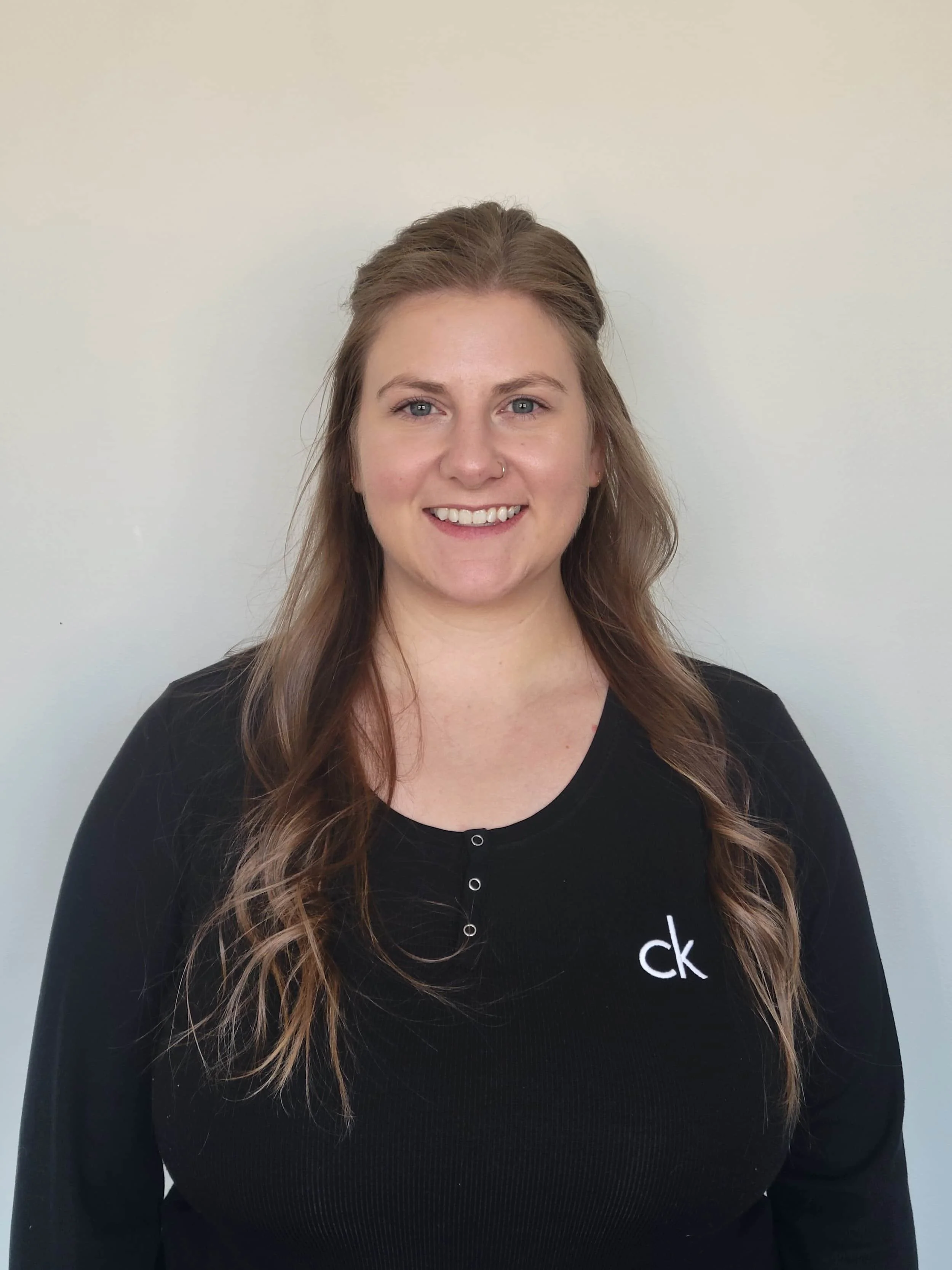 Woman with long, wavy brown hair smiles at the camera, wearing a black CK shirt—an osteopath standing against a plain light wall.