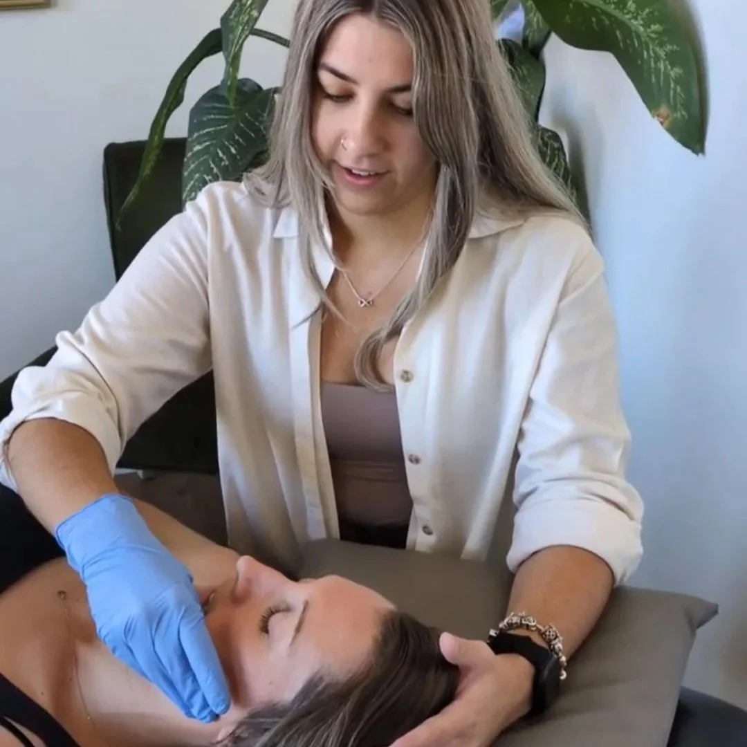 A woman in a white shirt gives a relaxing facial treatment for jaw pain relief TMJ to another woman lying down, wearing a blue glove.