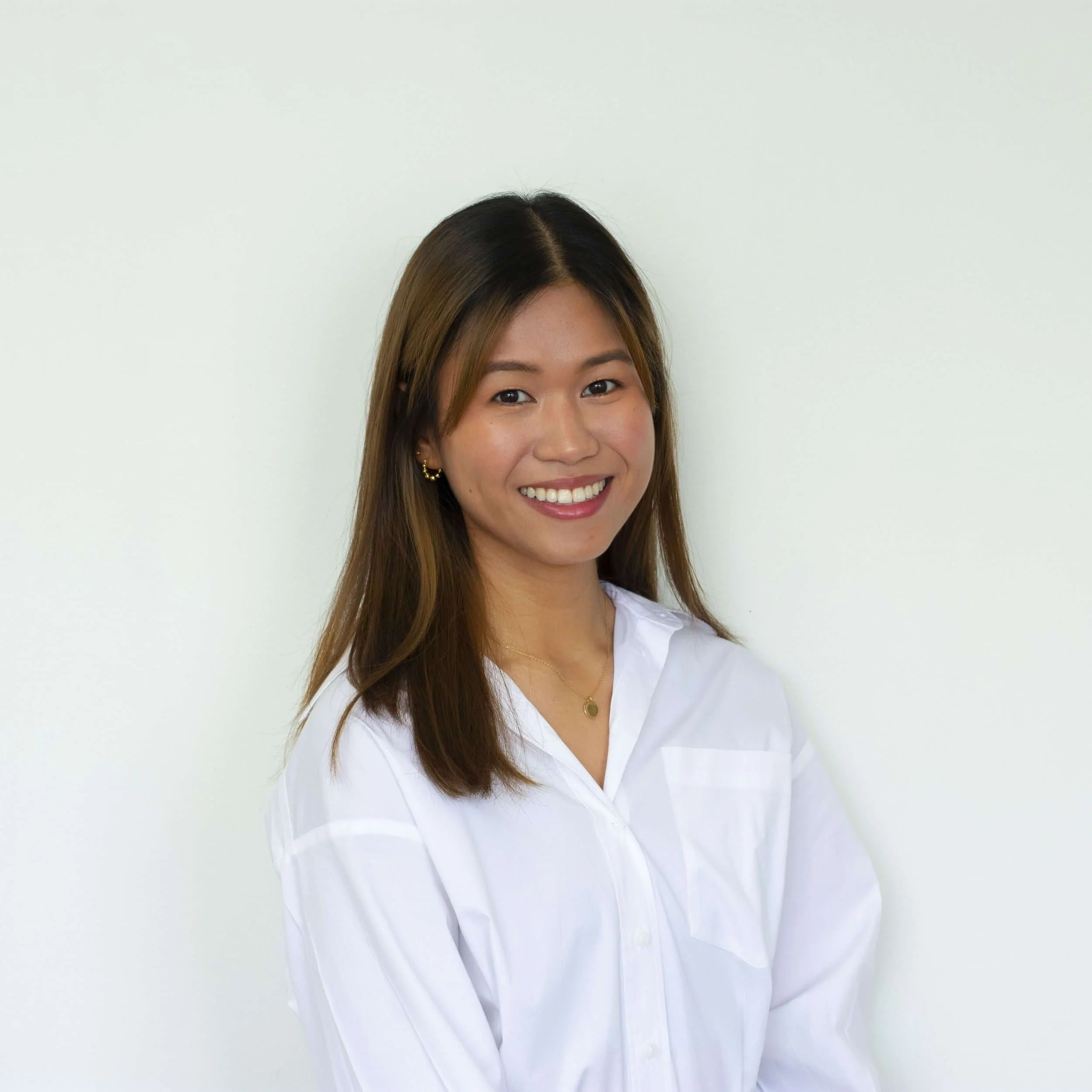 Woman with straight brown hair, wearing a white shirt, smiling, stands against a white background—learn about headache causes here.