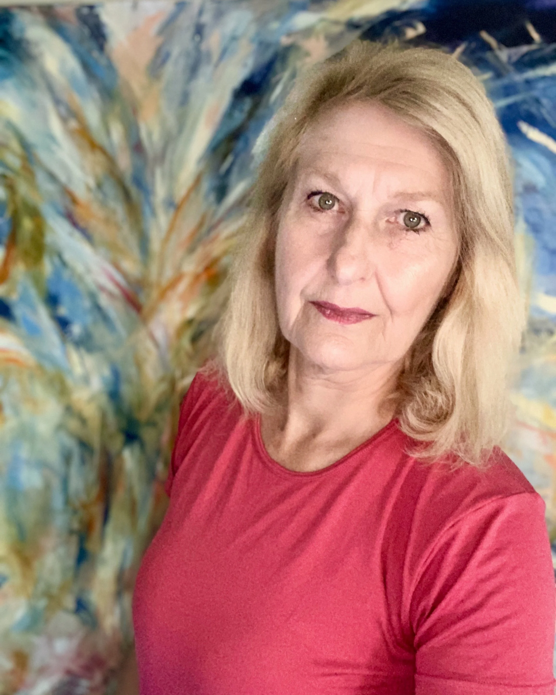 portrait of RM Cimini A woman with shoulder-length blonde hair taking a selfie in front of one of her abstract, colorful painting with shades of blue, green, yellow, and orange.