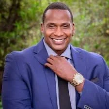 Smiling man in a blue suit and tie outdoors with greenery background