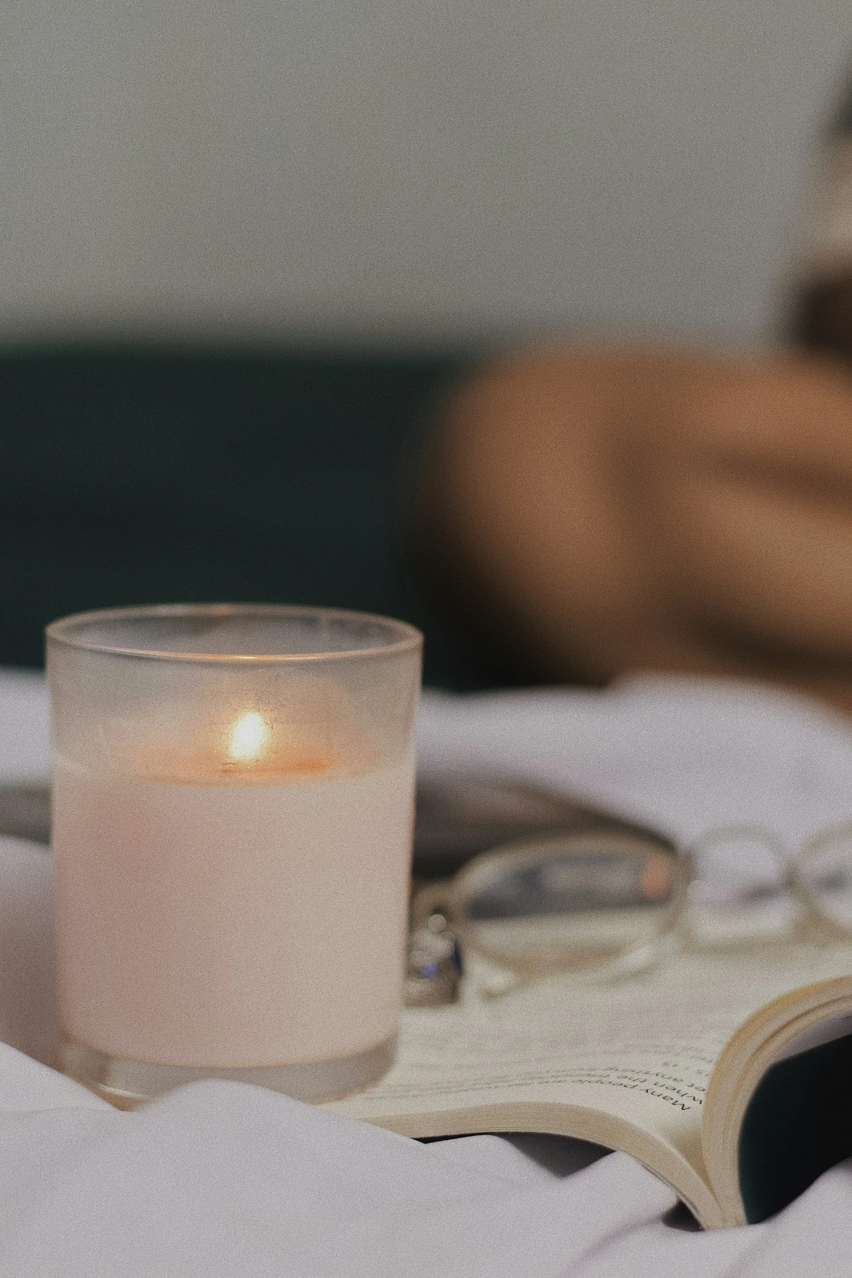 A lit candle, pair of eyeglasses, an open book with visible text, and a blurred person in the background.