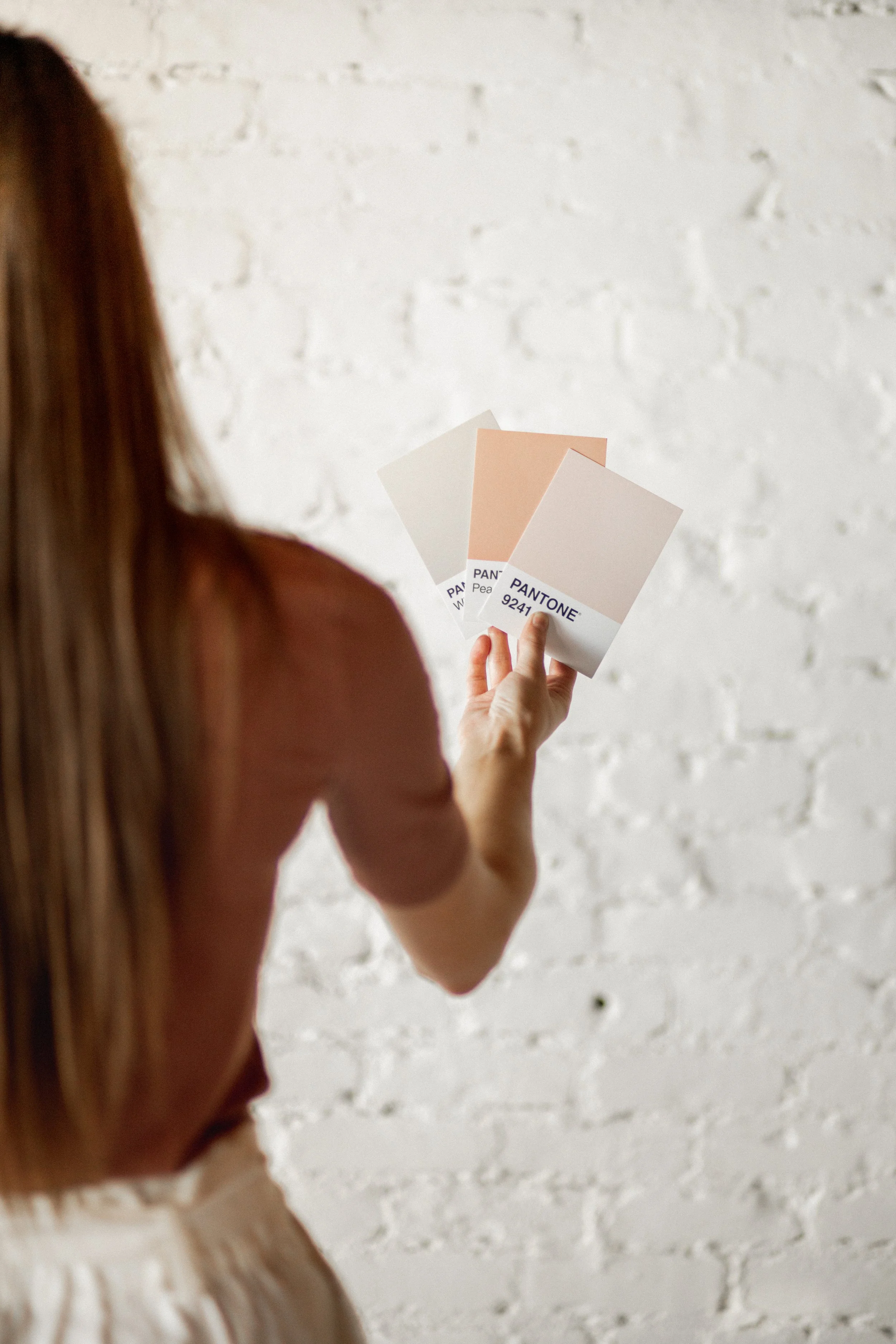 A woman holding color swatches or paint samples, including shades labeled 'Pantone 9241', in front of a white brick wall.