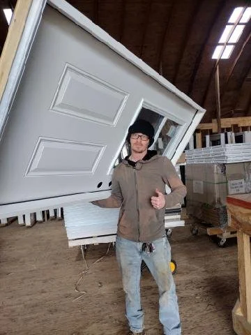 A man wearing a black beanie, glasses, and a brown jacket giving a thumbs up inside an industrial warehouse with a large tilted white garage door.