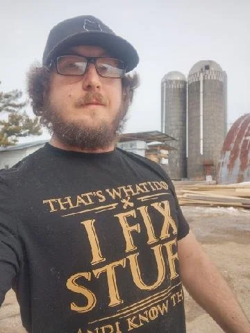 A man with glasses and a beard wearing a black baseball cap and a T-shirt that says 'That's what I do, I fix stuff, I know how to'. He is outdoors with two tall silos and a red barn in the background.
