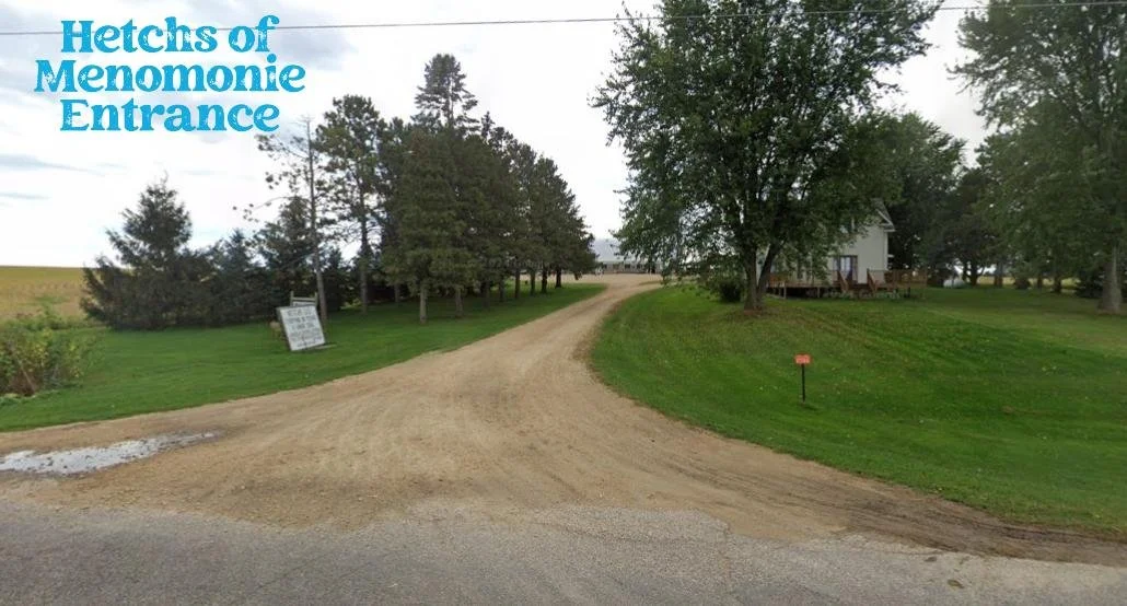 View of the entrance to Hetchs of Menomonie, featuring a gravel driveway winding through a grassy area with trees, near a white house on the right, and a sign on the left.