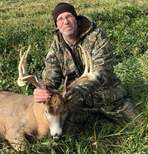 A man dressed in camouflage clothing and a black beanie kneeling in tall grass, holding the antlers of a large deer with antlers.