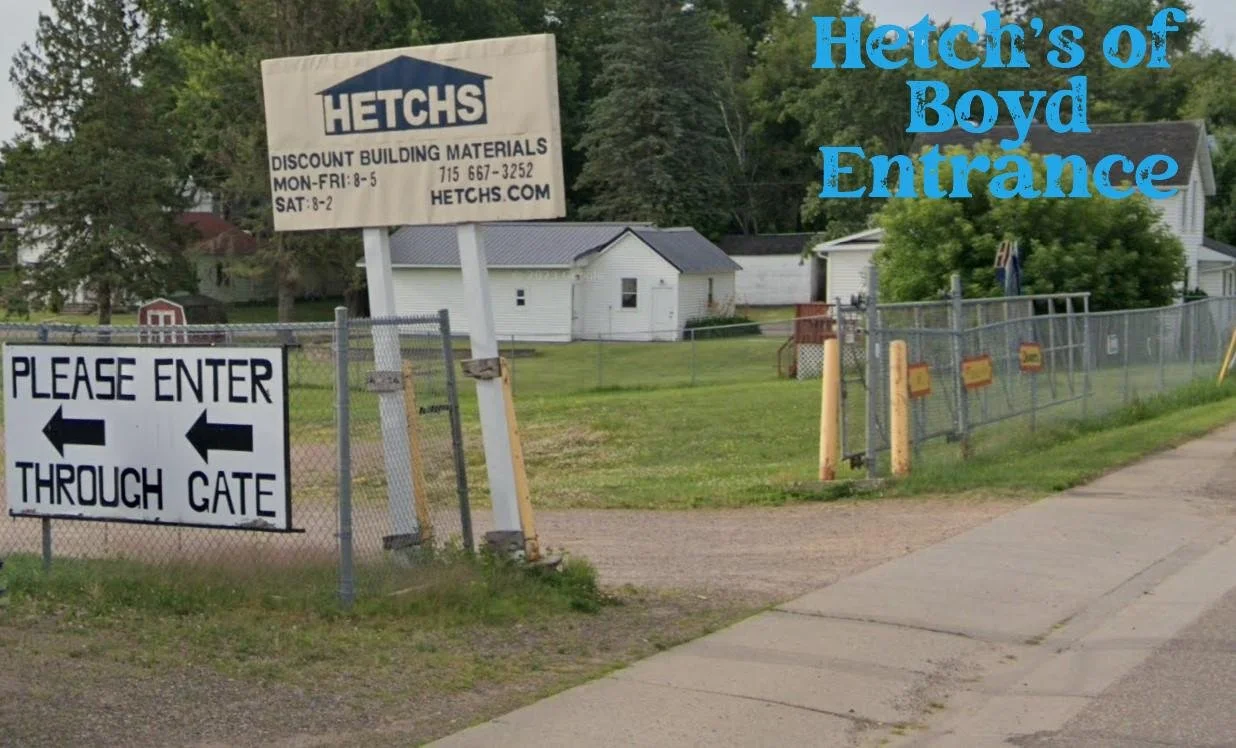 Entrance to Hertz's of Boyd with a sign that says 'Please Enter Through Gate' and a large Hetch's sign advertising discount building materials, located in a grassy area with houses in the background.