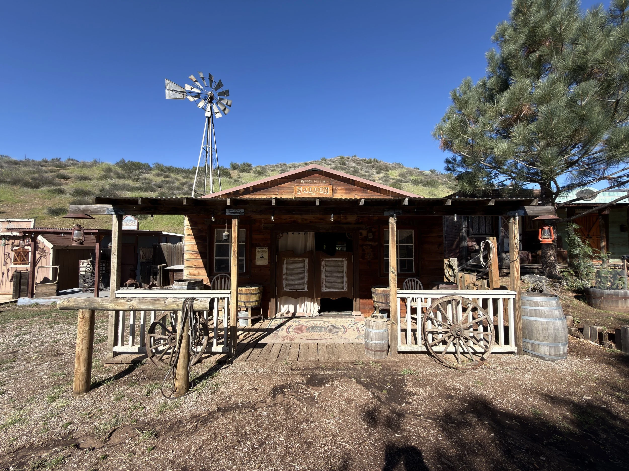 Location scouting photo of the standing Western saloon set at Agua Dulce used for On A Razor's Edge, showing the wooden facade, covered porch, and windmill under clear daytime skies.