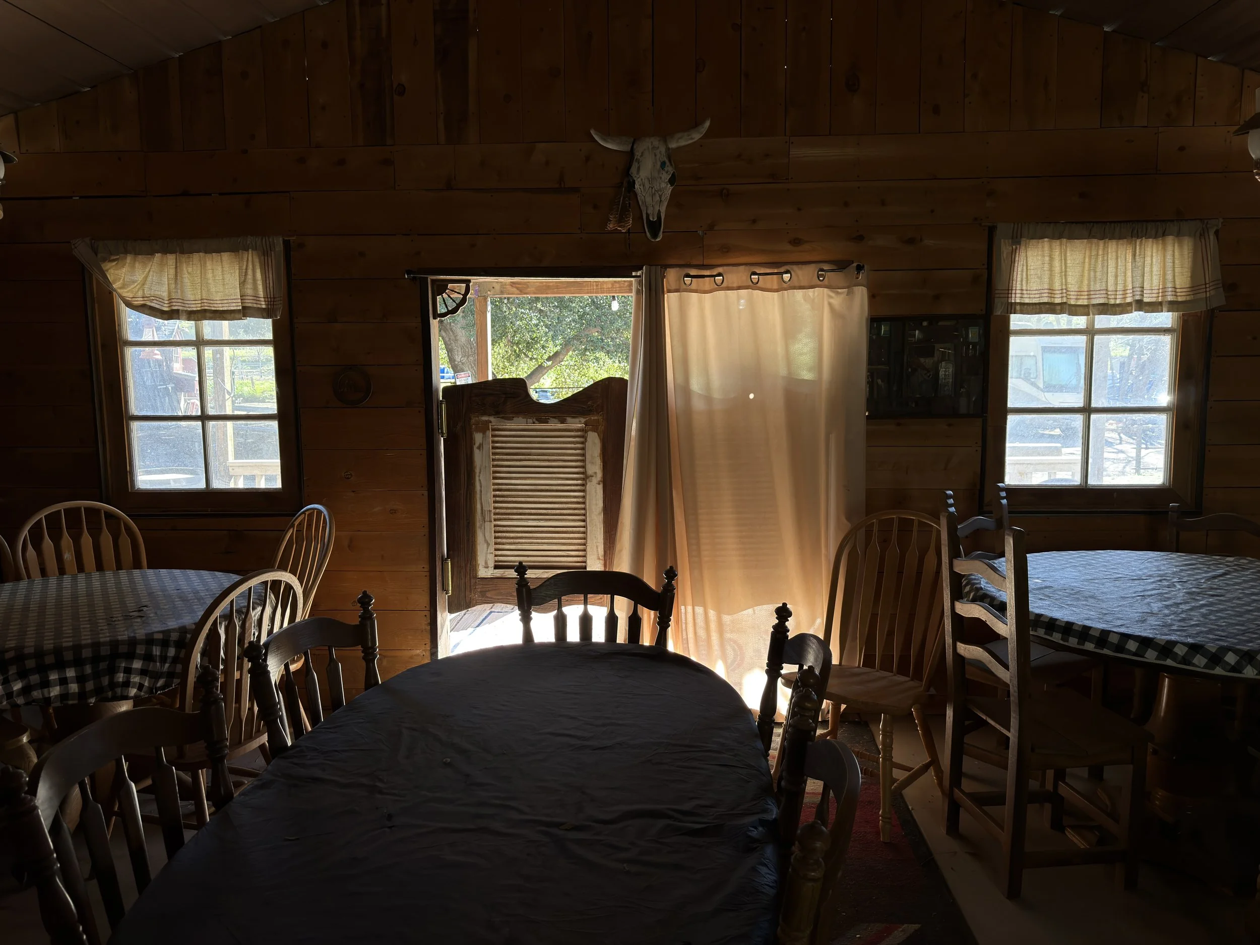 Location scouting photo of the On A Razor's Edge saloon set dining area, with checkered tablecloths, a longhorn skull mount, and bright daylight streaming through the open doorway.