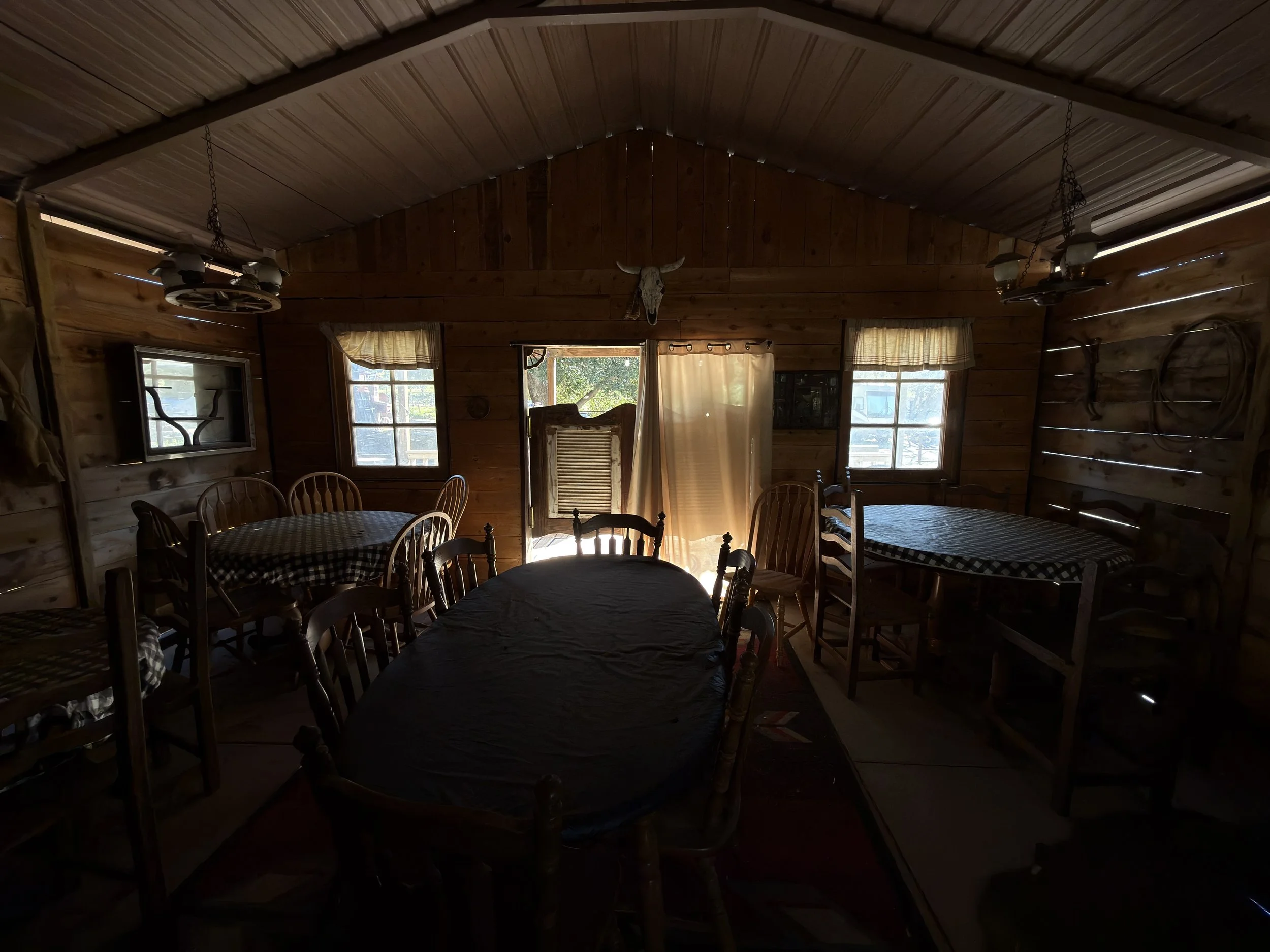 Wide-angle location scouting photo of the On A Razor's Edge saloon set showing the vaulted wood ceiling, dining tables, string lights, and sunlit doorway before the production's rainy shoot day.
