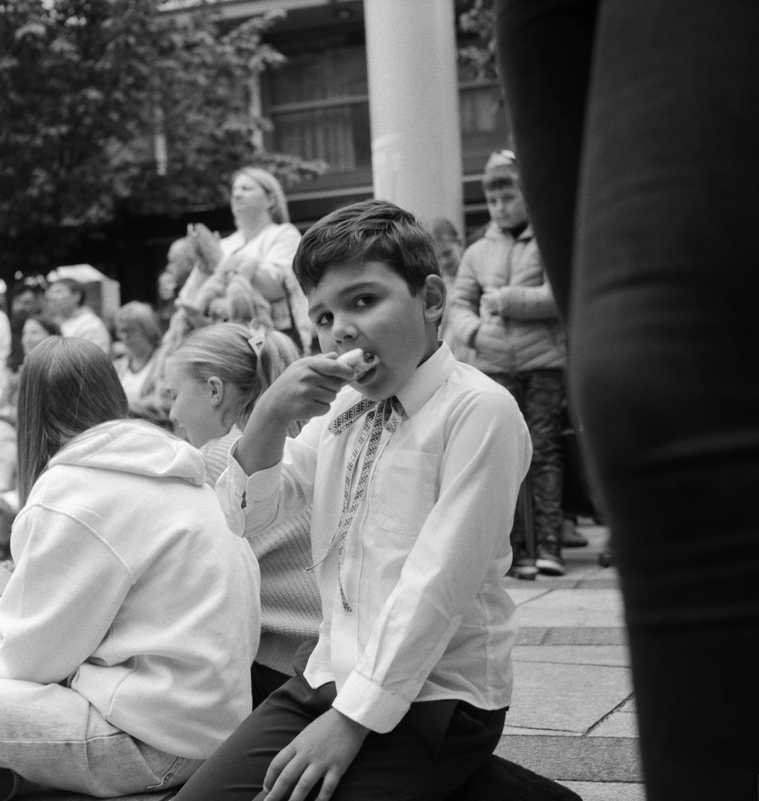 A young boy with dark hair sitting on the ground, eating something, during a gathering or event with other children and adults in the background.