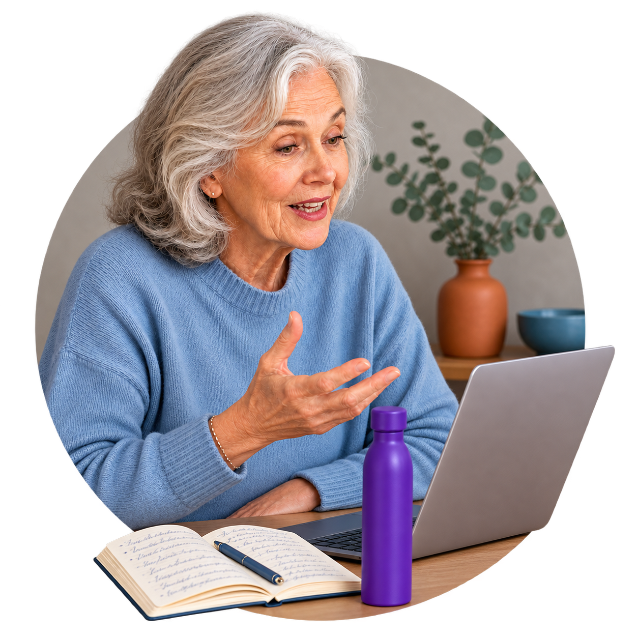Older woman with gray hair wearing a blue sweater talking during a video call, sitting at a desk with a laptop, open notebook, purple water bottle, and a pen.