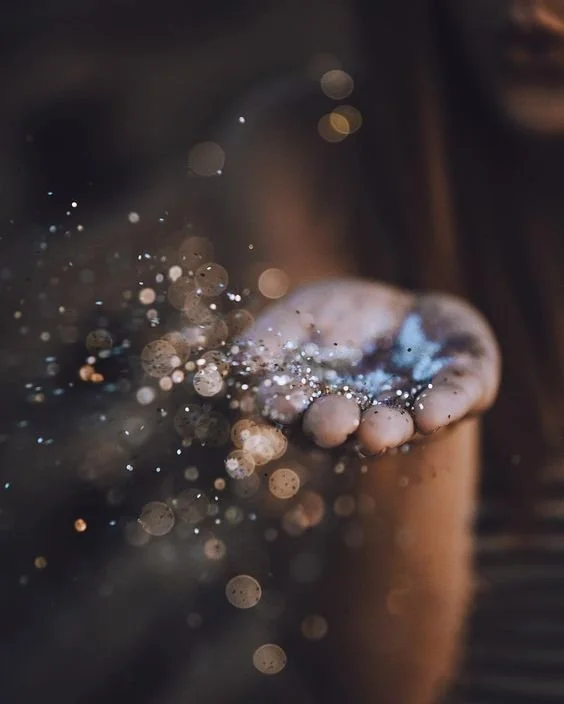 Close-up of a person's hand holding a handful of sparkles or glitter, with light reflecting off them creating a bokeh effect.