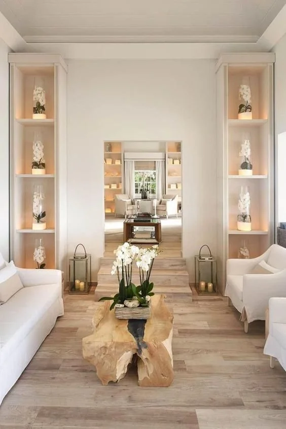 Living room with sofas, a wooden coffee table with orchids, built-in shelves with white decorative objects, and lanterns with candles on the floor. A doorway leads to a bright sitting area with armchairs and a window. An example of good Feng Shui.