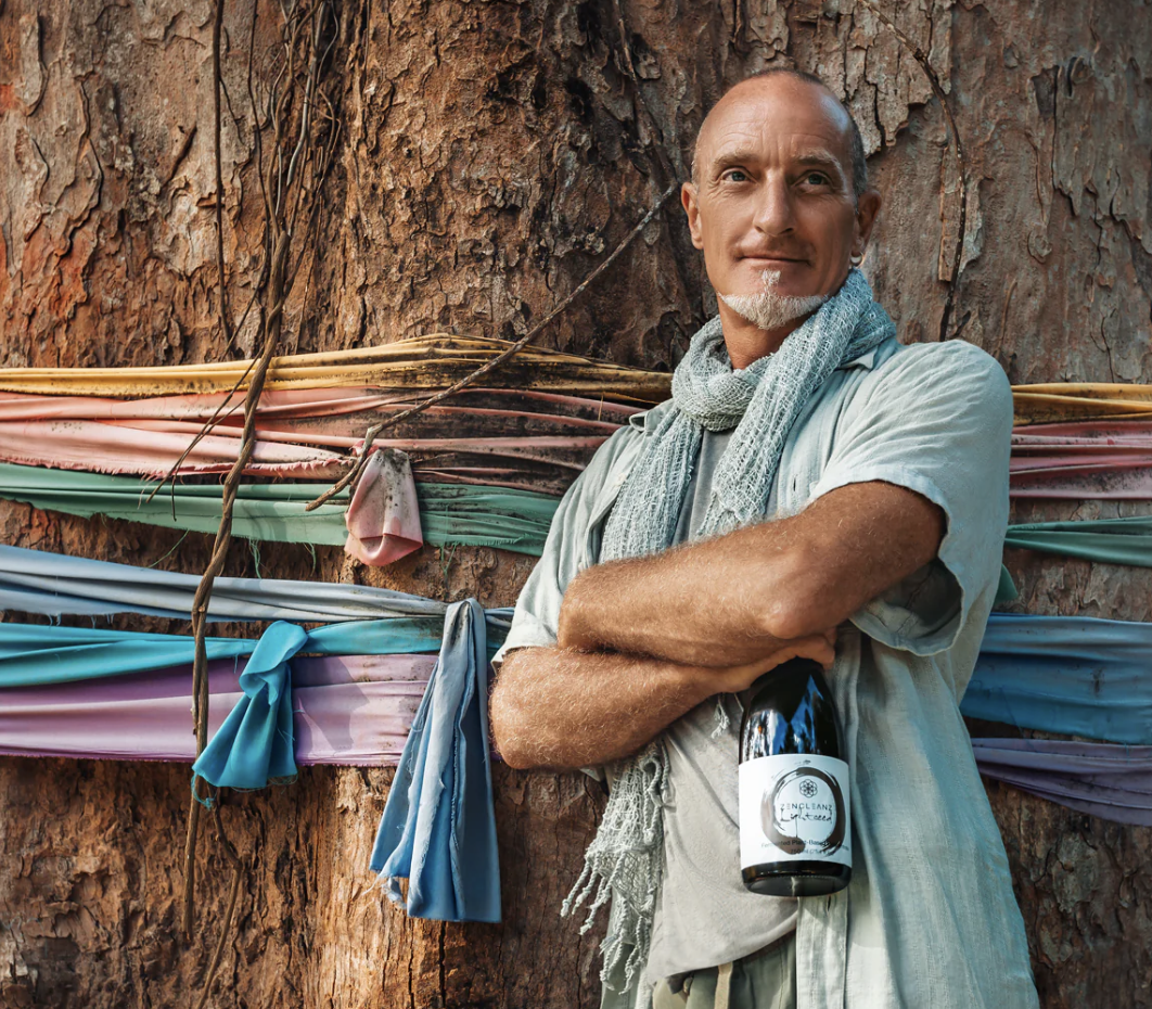 A man with a scarf around his neck, standing next to a large tree with colorful cloth wrapped around it, holding a bottle in his hand. He is a ZenCleanz affiliate partner.