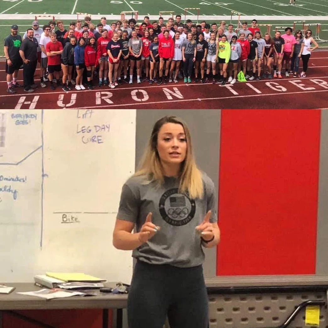Group of people on a football field, posing for a photo, with 'HURON TIGERS' written on the track. A woman with blonde hair, wearing a gray T-shirt, stands in front of a whiteboard with notes, pointing with her index fingers.