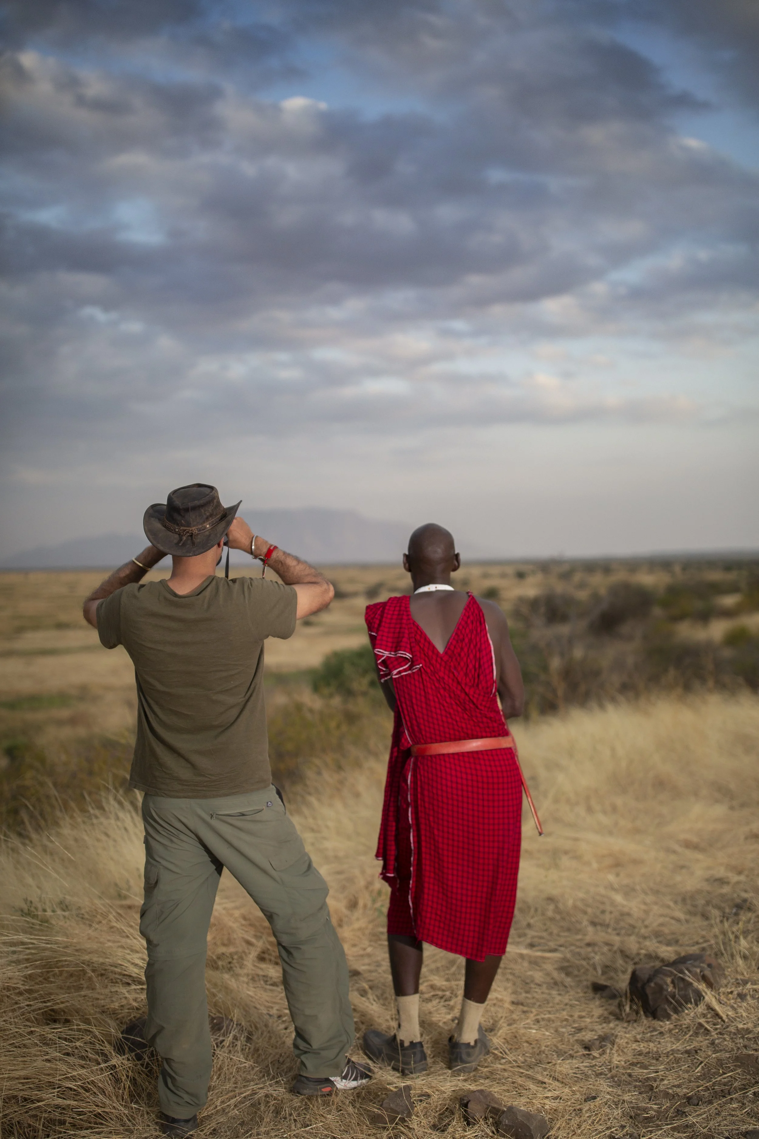 Two men, one in casual clothing and a hat, and the other in traditional Maasai attire, stand in an open, grassy landscape under a cloudy sky, observing the horizon.