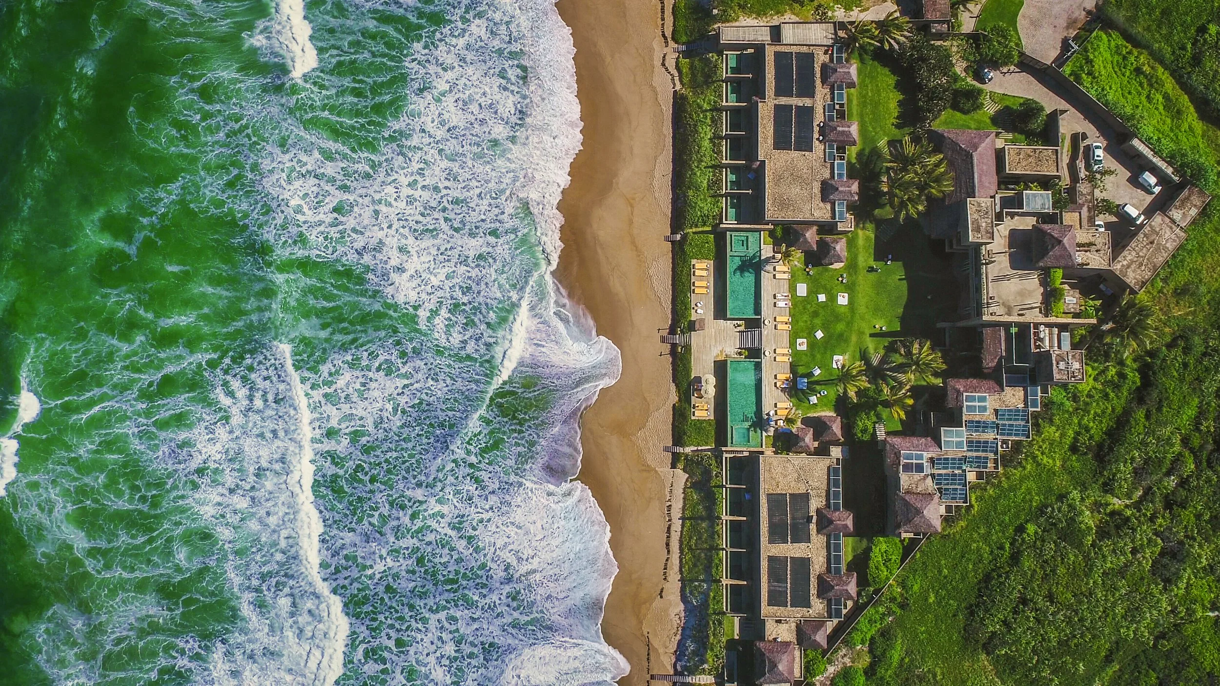 Aerial view of a beachside resort with swimming pools, lounge chairs, and a large building surrounded by greenery, overlooking the ocean with waves crashing onto the sandy shore.