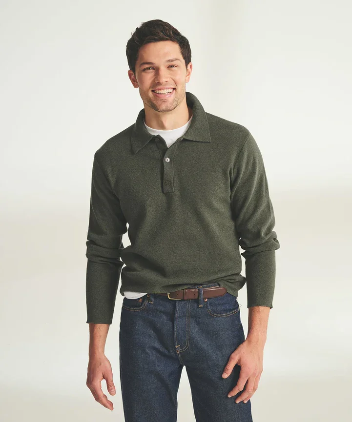 A young man smiling, wearing a dark green sweater over a white shirt and blue jeans, standing against a plain light background.