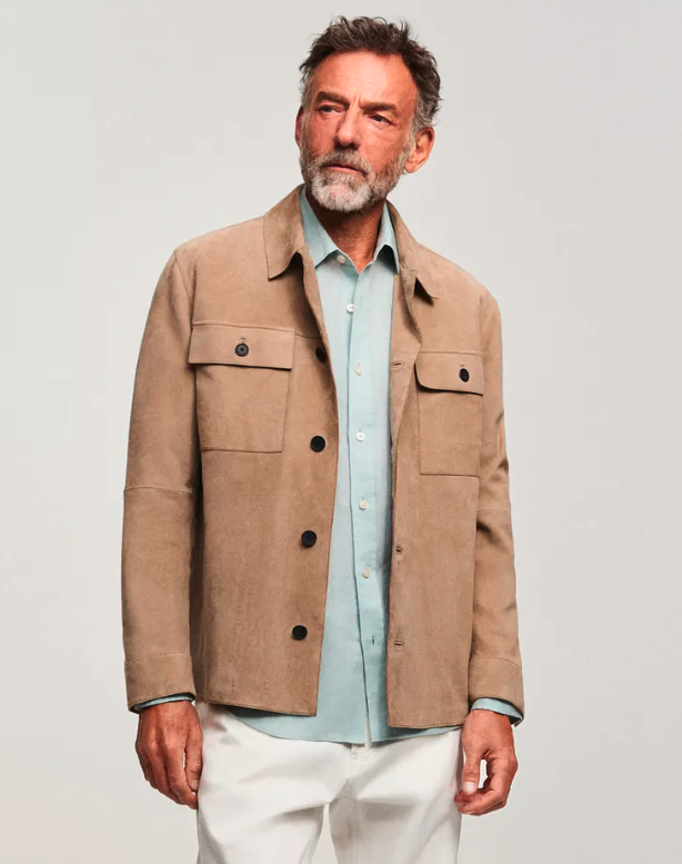 An older man with gray hair and beard wearing a beige jacket over a light blue button-up shirt and white pants, standing against a plain light background.