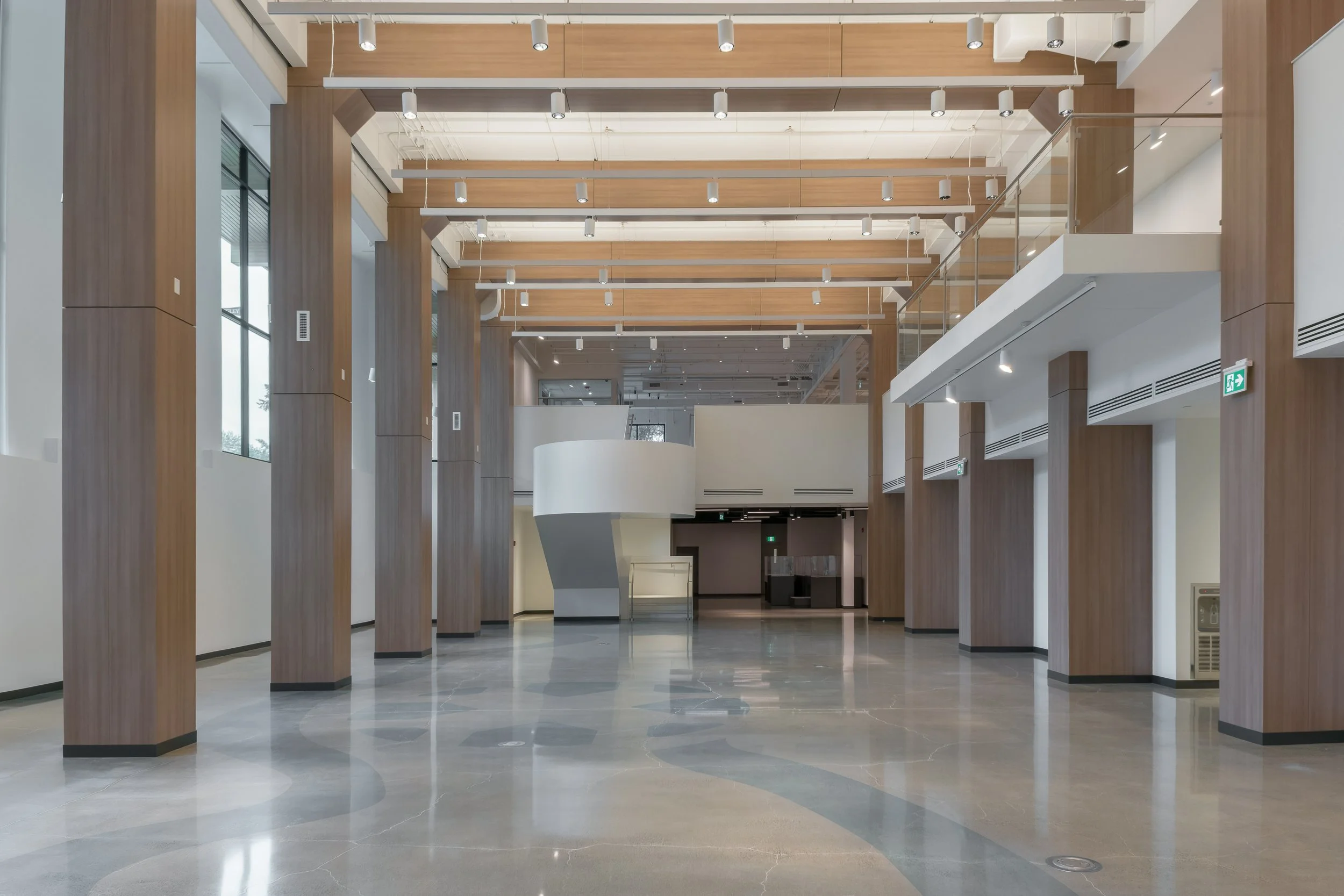 Empty interior space with tall wooden columns, modern lighting, and a staircase in the center, featuring a polished marble floor and glass railings.