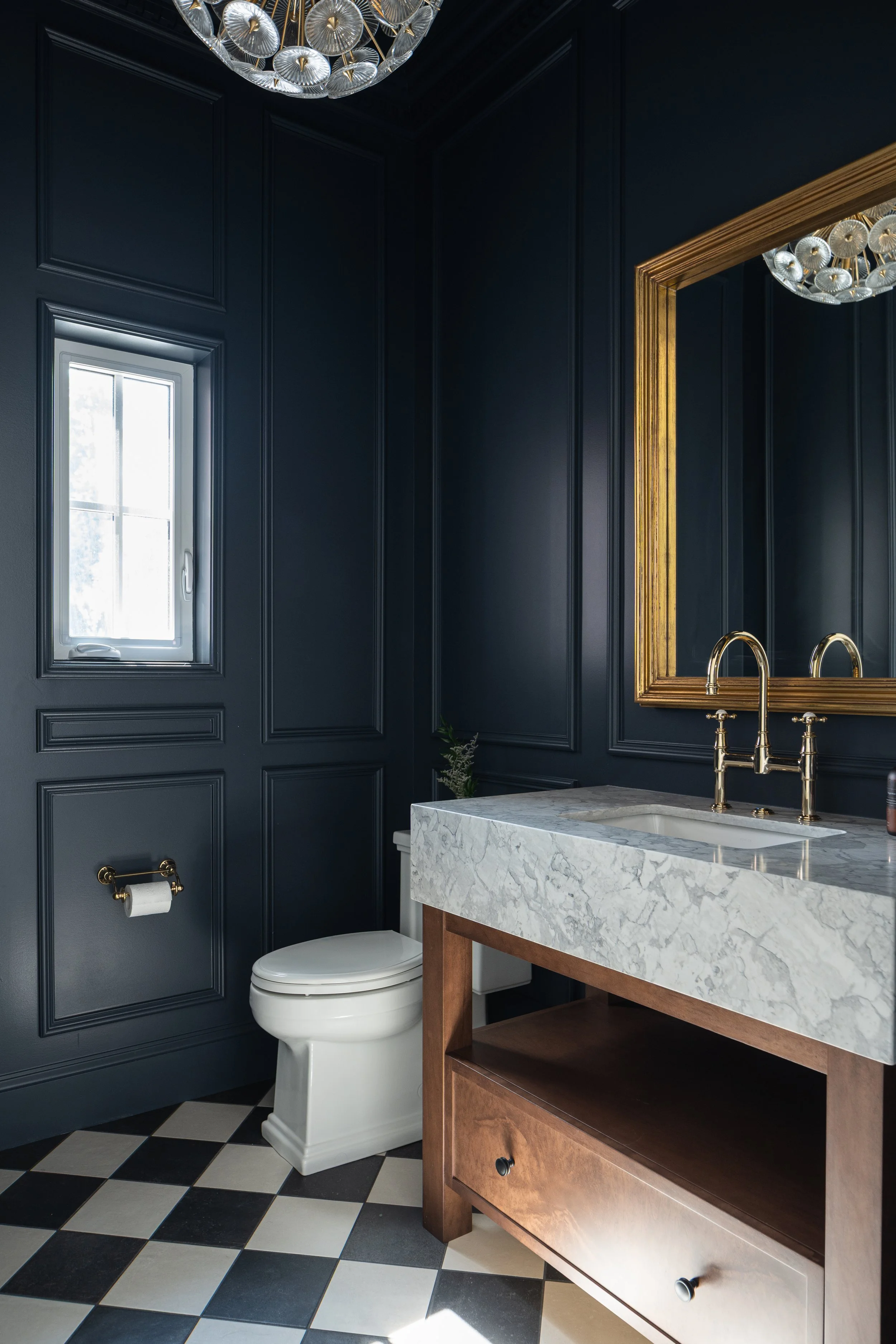 Interior design of a A luxurious bathroom with dark blue paneled walls, a window, a white toilet, a marble vanity with a gold faucet, a large gold-framed mirror, and a black and white checkered floor.
