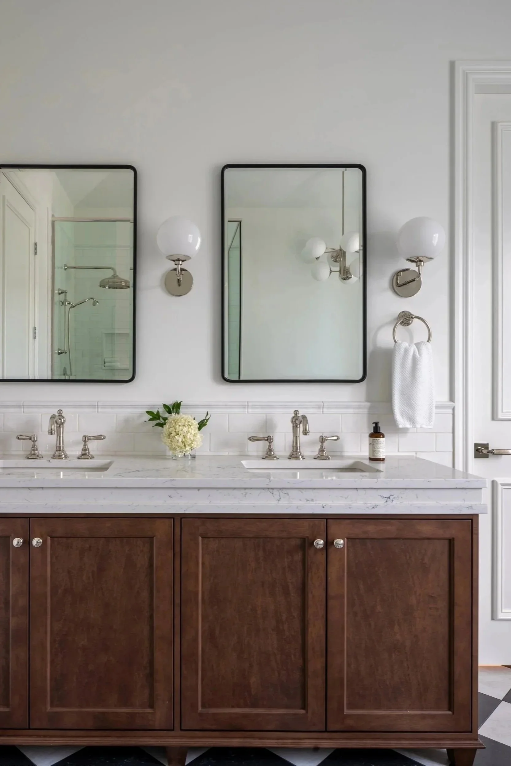 Interior design of a A bathroom vanity with a marble countertop, two sinks, and two mirrors, flanked by wall-mounted sconces and a towel ring. There is a flower vase and a soap dispenser on the counter.