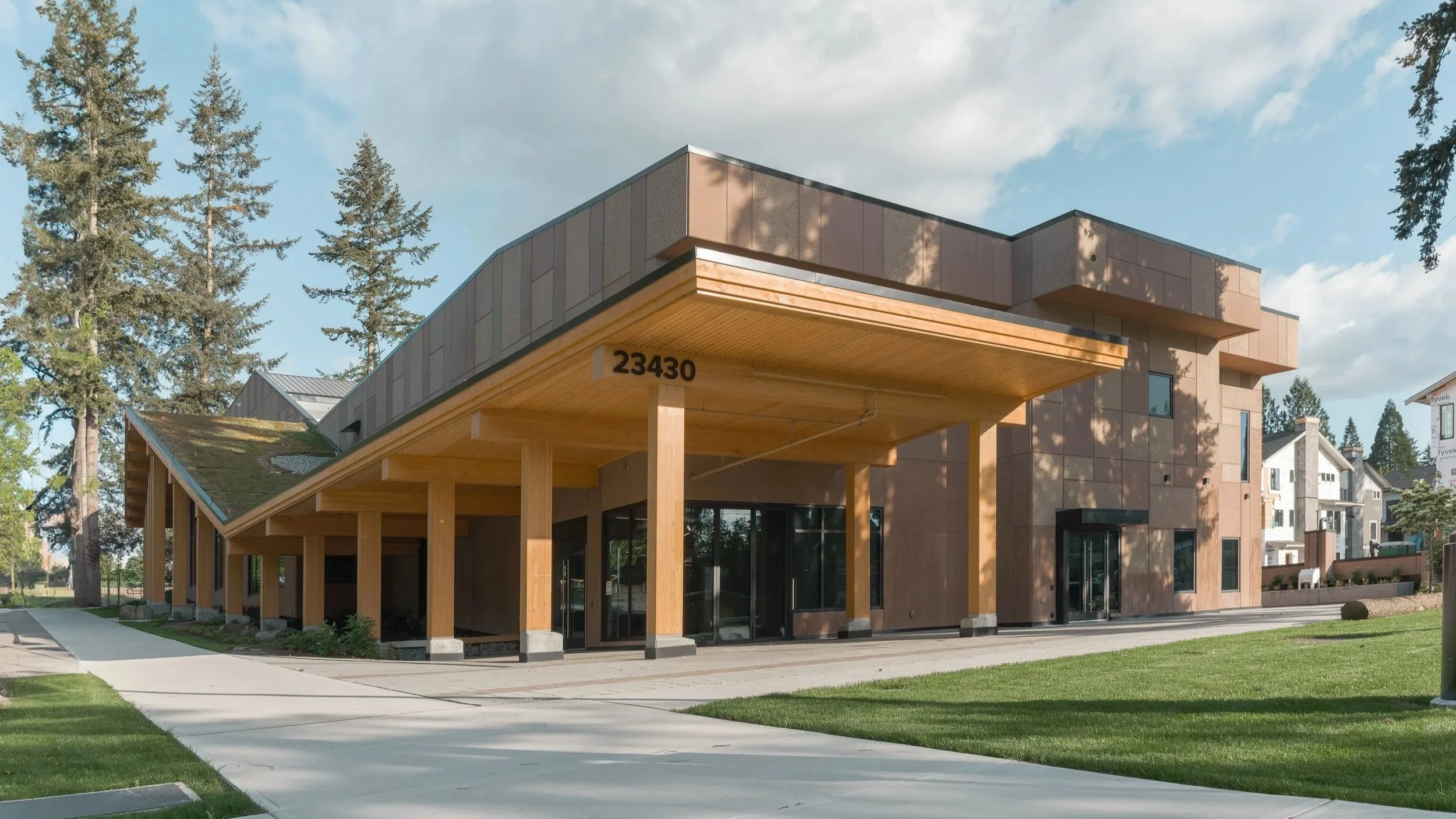 Modern building with a combination of wood and concrete exterior, tall glass windows, and a covered entrance supported by wooden columns, with a grassy lawn and sidewalk in front.