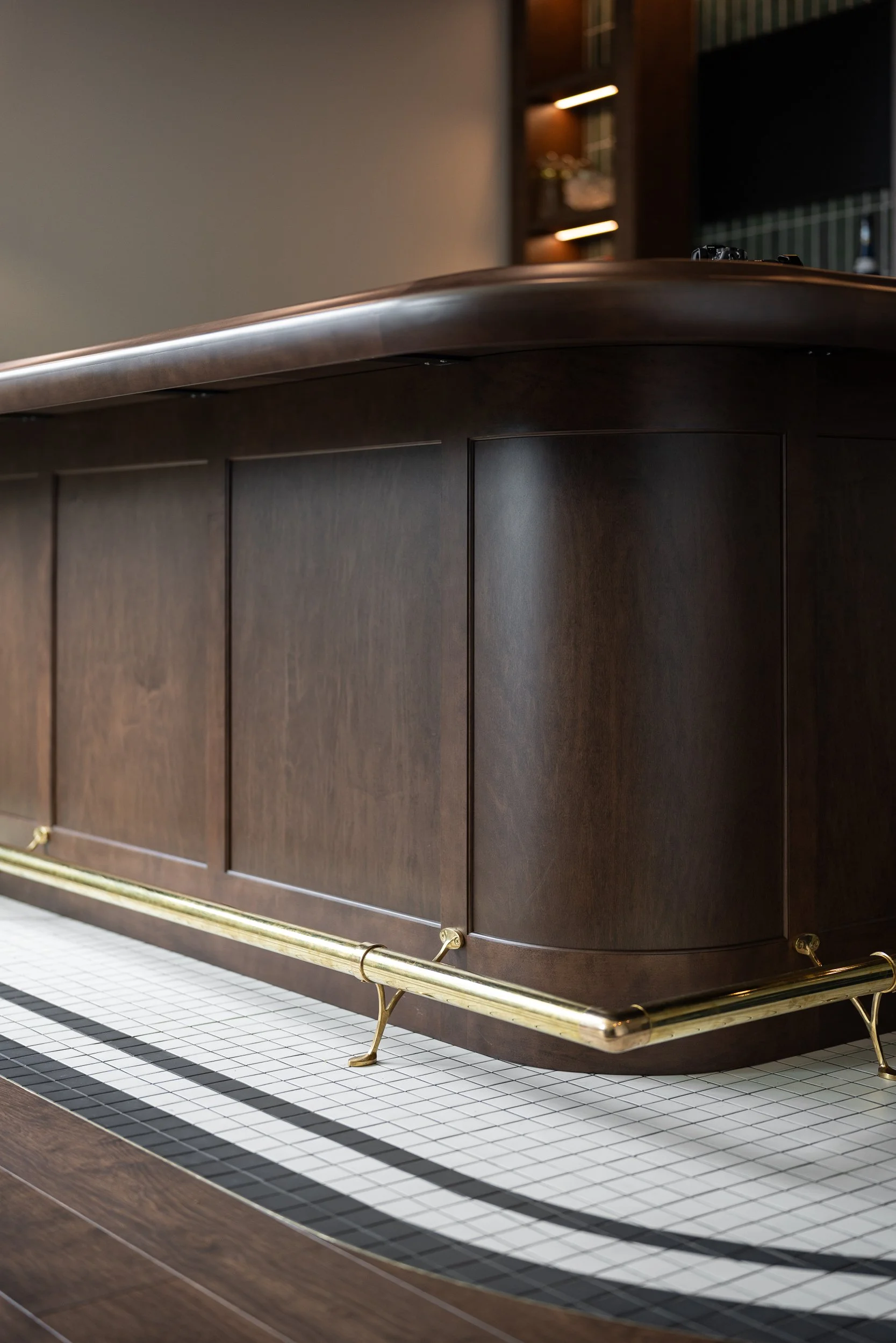 Interior design of a Close-up of a dark wooden bar with rounded edges, brass foot rail, and a tiled floor.