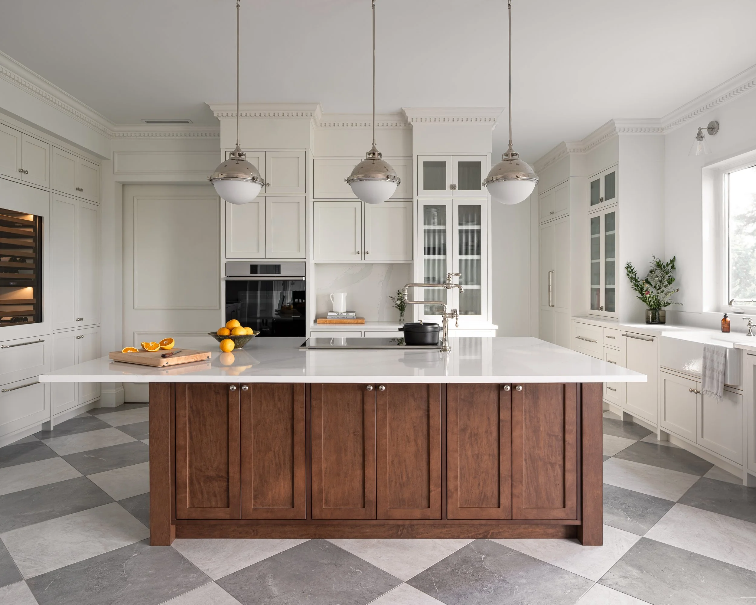 Interior design of a modern kitchen with white cabinets, a large white central island with wooden base, checkered gray and white tile floor, hanging pendant lights, window with plant, and lemons on the island countertop.