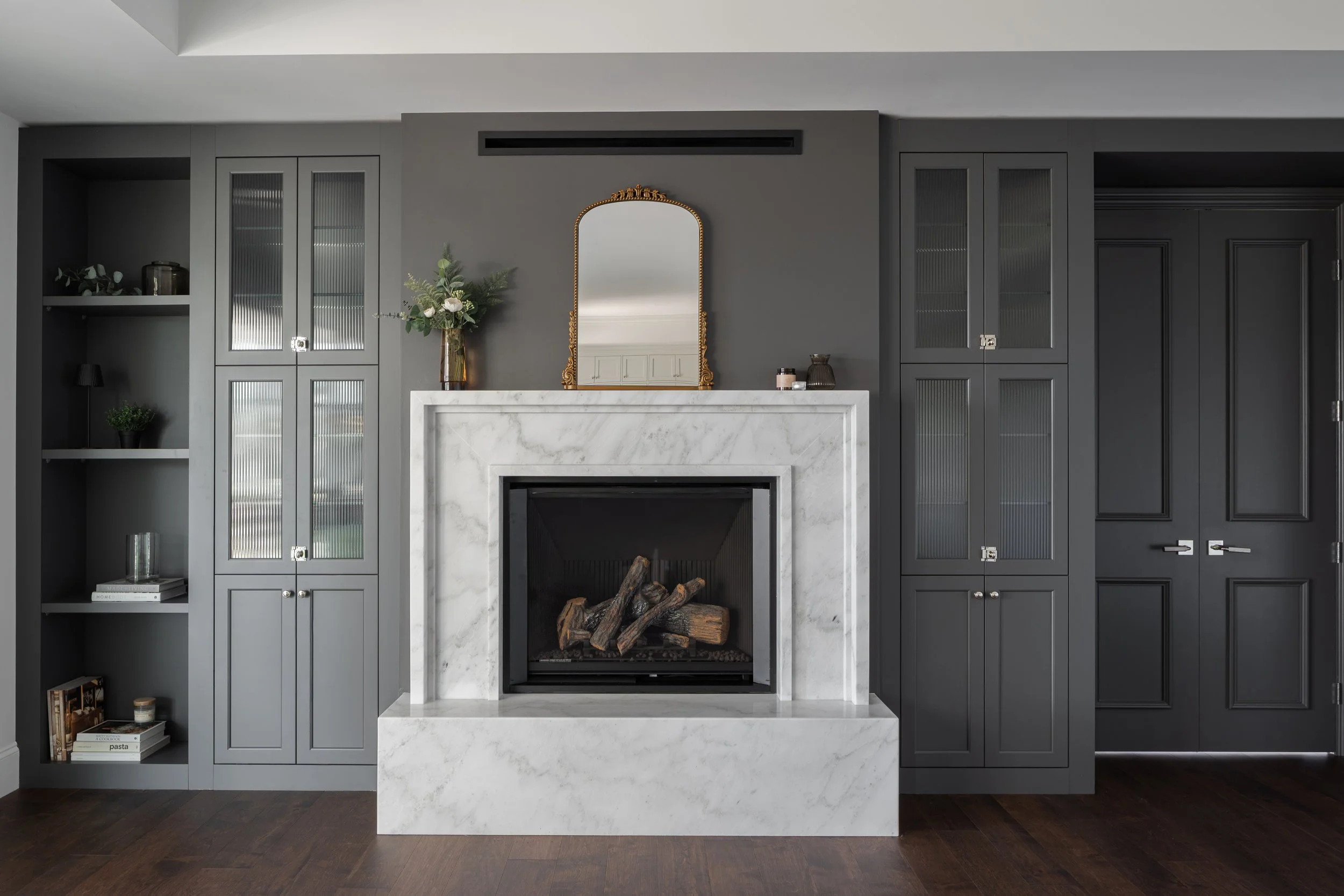 Interior design of a Living room with a modern white marble fireplace, dark gray built-in cabinets on both sides, and a gold-framed mirror hanging above the fireplace.