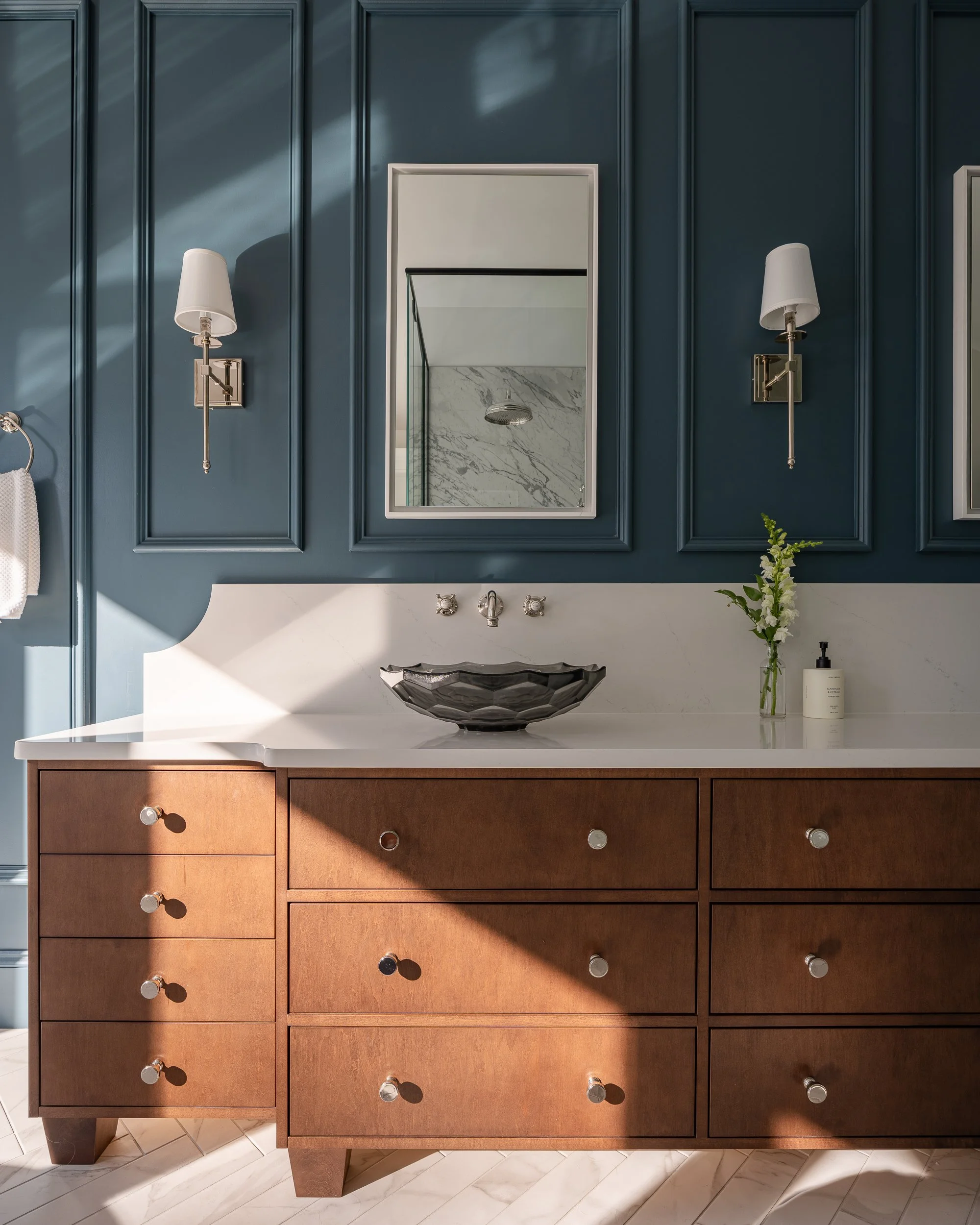 Interior Bathroom vanity with a brown wooden cabinet, a white countertop, a black vessel sink, and a rectangular mirror. Two wall-mounted lamps are on either side of the mirror. Blue paneled walls and a marble shower wall are reflected in the mirror.